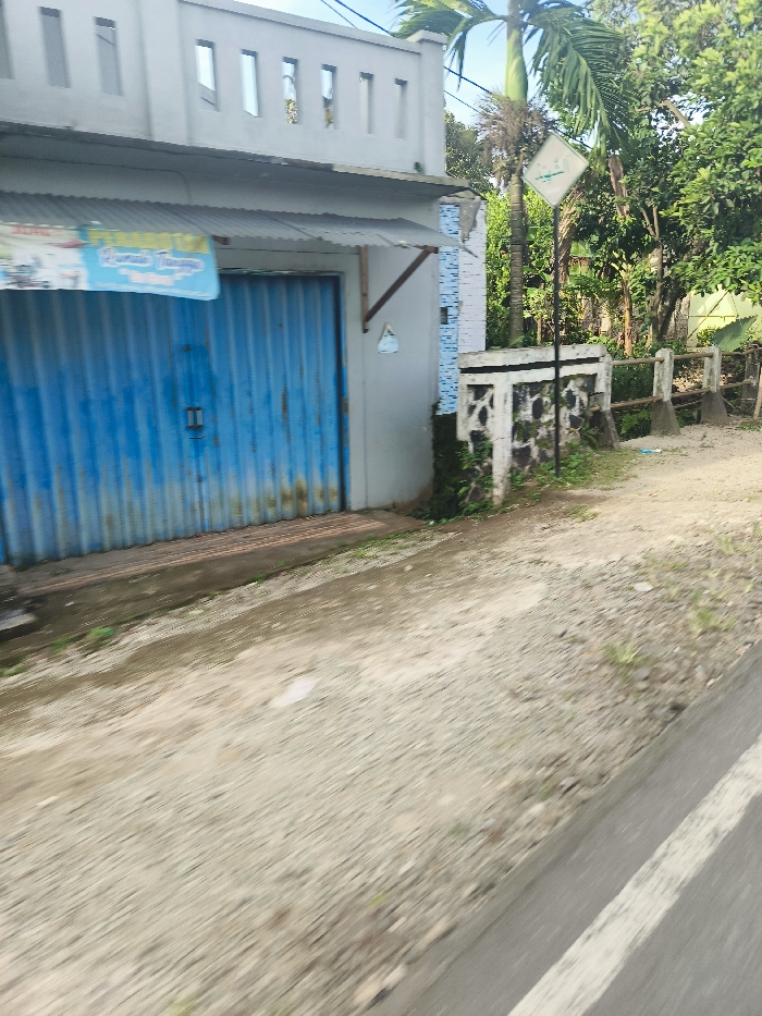 The image shows a blue metal roll-up door on the side of a building in Sukatani, Indonesia, located at coordinates -6.643 latitude and 107.414 longitude. The building is grey with a white trim, and the door is partially open. The building is surrounded by a small stone wall. The door is situated in front of a gravel road that extends away from the viewer, and a white painted line runs along the edge of the road. There are no people visible in the image, but there is greenery and vegetation surrounding the building. A road sign can be seen in the background, but the image is not close enough to read the sign. The image was likely taken by someone passing by on the road in a vehicle.