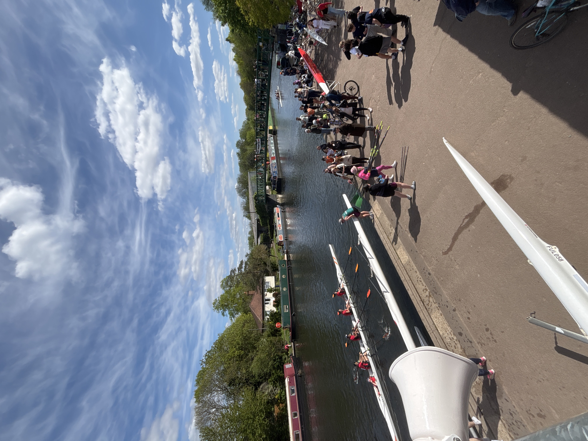 The image depicts a rowing event on a river, possibly the River Thames in London, United Kingdom, given the urban and architectural elements visible. A crowd of spectators lines the bank, observing the rowing crews in their boats. Several rowing boats, each with multiple rowers, are visible in the water. The sky is partly cloudy, and buildings and trees line the riverbank. A loudspeaker is visible in the foreground. People are standing on a concrete path near the river, some holding oars.