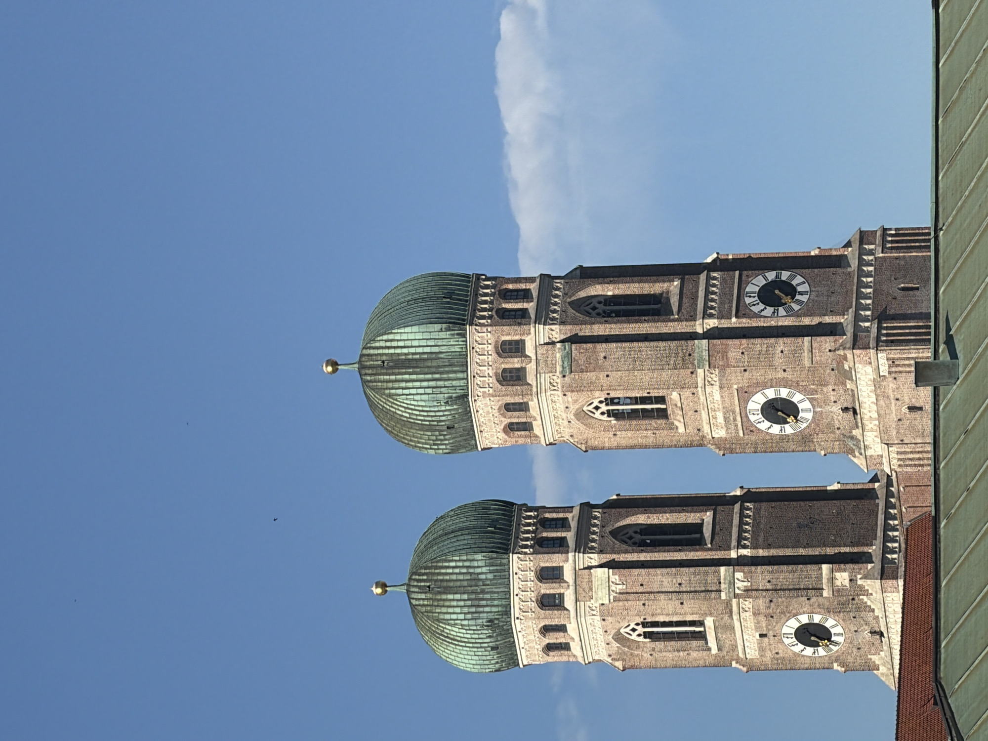 This is a close-up, eye-level, outdoor, daytime, medium shot of two identical, tall, brick church towers with green, onion-shaped domes topped with gold finials. The towers have Gothic architectural details, including arched windows and decorative stonework. Each tower has a large clock face with Roman numerals and black hands. The sky behind the towers is a clear, bright blue with a single, thin, wispy white cloud on the right side. A few small, dark specks, possibly birds, are visible in the sky. The tops of two buildings with green and red tile roofs are visible in the foreground, partially obscuring the bases of the towers. The image captures the Frauenkirche (Church of Our Lady) in Munich, Germany.