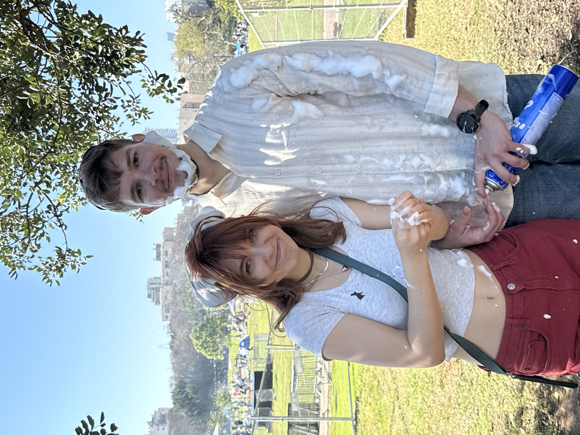 This outdoor image, rotated 90 degrees clockwise, captures two young adults, a man and a woman, engaged in a playful foam fight on a bright, sunny day in what appears to be a park or event space in Tel-Aviv, Israel.
The man, positioned slightly behind and above the woman, has fair skin, short dark hair, and stubble on his face. He is wearing a light-colored, long-sleeved collared shirt and dark pants. His face, neck, and shirt are generously covered in white foam. He holds a blue aerosol can, partially obscured by his hand, with fragmented white text visible, possibly indicating its contents. A dark wristband is on his left wrist. He looks towards the camera with a slight smile.
The woman, in the foreground, has fair skin, long reddish-brown hair with bangs, and is wearing a light gray t-shirt with a small dark embroidered emblem (resembling an animal) and red/maroon pants. Her midriff is exposed. Her hands, arms, and stomach are also splattered with white foam. She smiles warmly at the camera. She wears a silver necklace and a choker, and a dark green strap (likely from a bag) crosses her body.
The background features a large grassy area, indicative of a park or an event ground. Metal barriers and distant crowds suggest a public gathering or festival. Beyond these, urban buildings are visible against a clear blue sky. Green tree branches with leaves frame the upper left and lower right corners of the image. The overall mood is joyful and energetic, with both subjects clearly enjoying the activity.