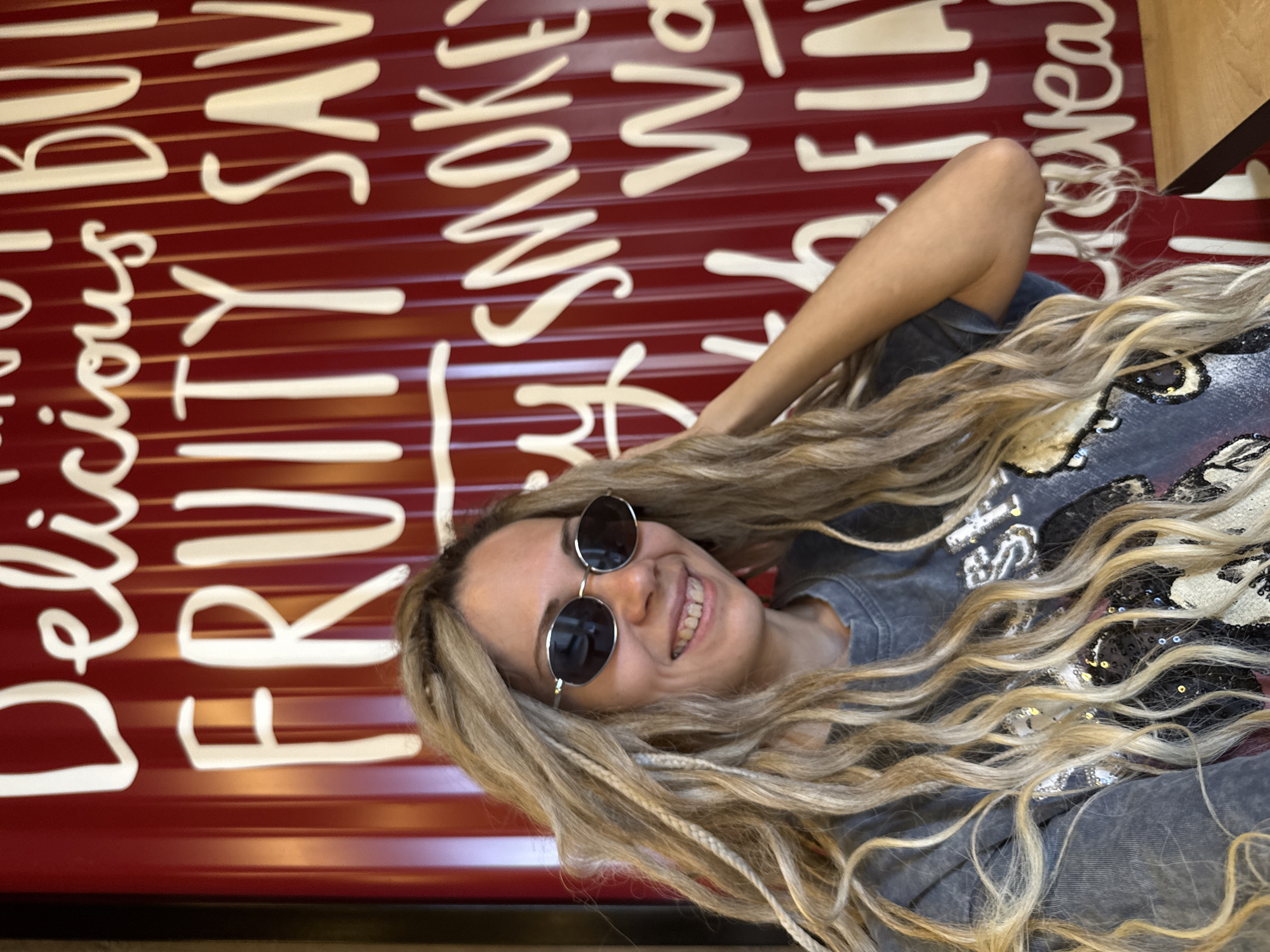 A woman with long blonde hair, braided in some sections, wears round, dark sunglasses and a grey t-shirt adorned with sequins. She smiles broadly, revealing her teeth, as she touches her hair with her right hand. The woman is positioned in front of a large, red wall with white lettering. The visible text on the wall reads "Delicious BU", "FRUITY SAV", "SMOKEY Wa", and "FLAV". The environment suggests an indoor setting, possibly a restaurant or cafe. The image does not contain any information to confirm the location in Шебунино, Russia.