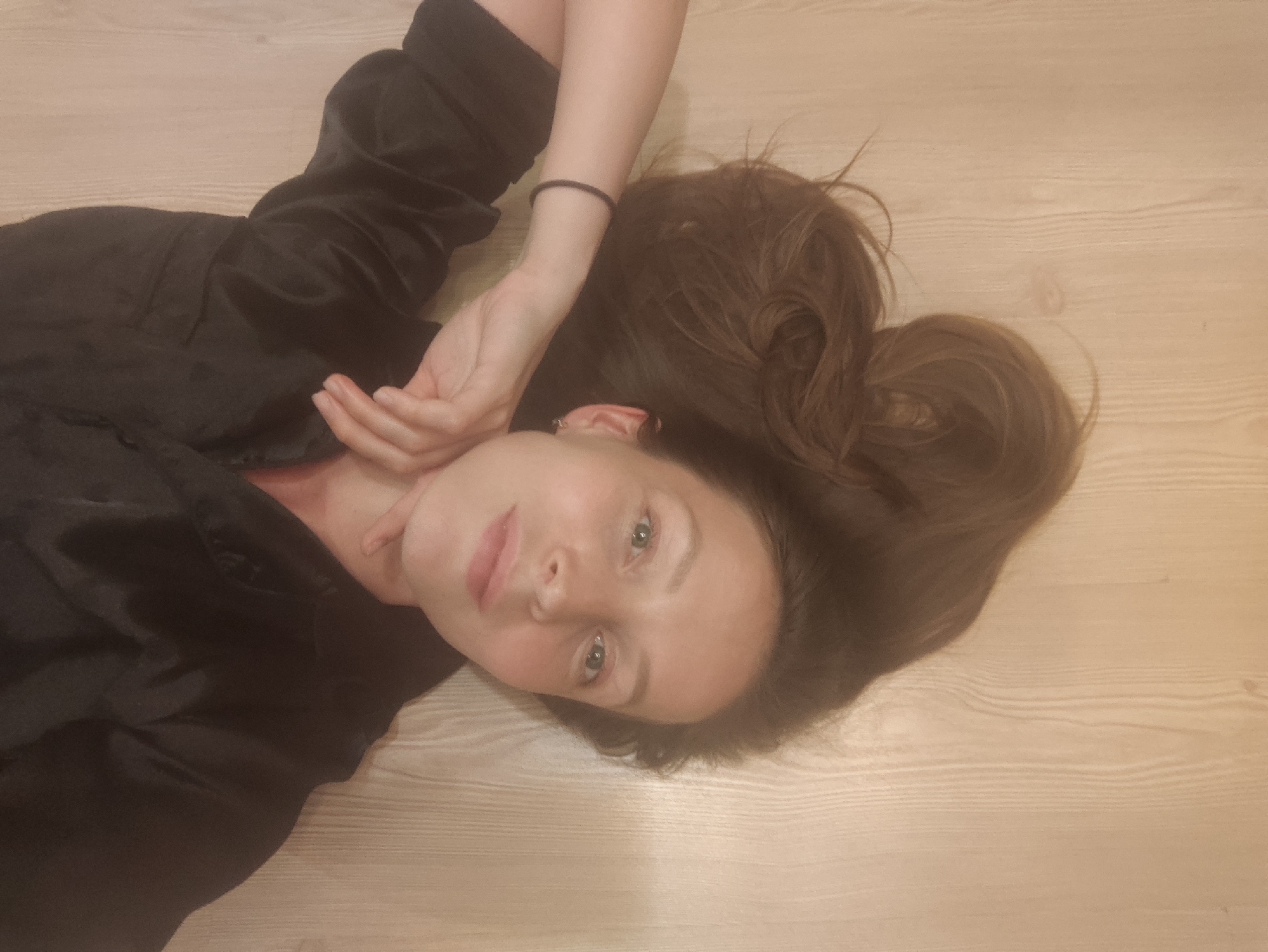 A woman lies on a light-colored, wooden floor. She is wearing a black satin-like garment with long sleeves, and her brown hair is spread out around her head. Her right hand is near her face, resting on her neck. A black hair tie is visible on her right wrist. Her face is tilted slightly upwards, and she is looking directly at the camera. The exact location in the city of Chaikovsky, Russia cannot be determined.