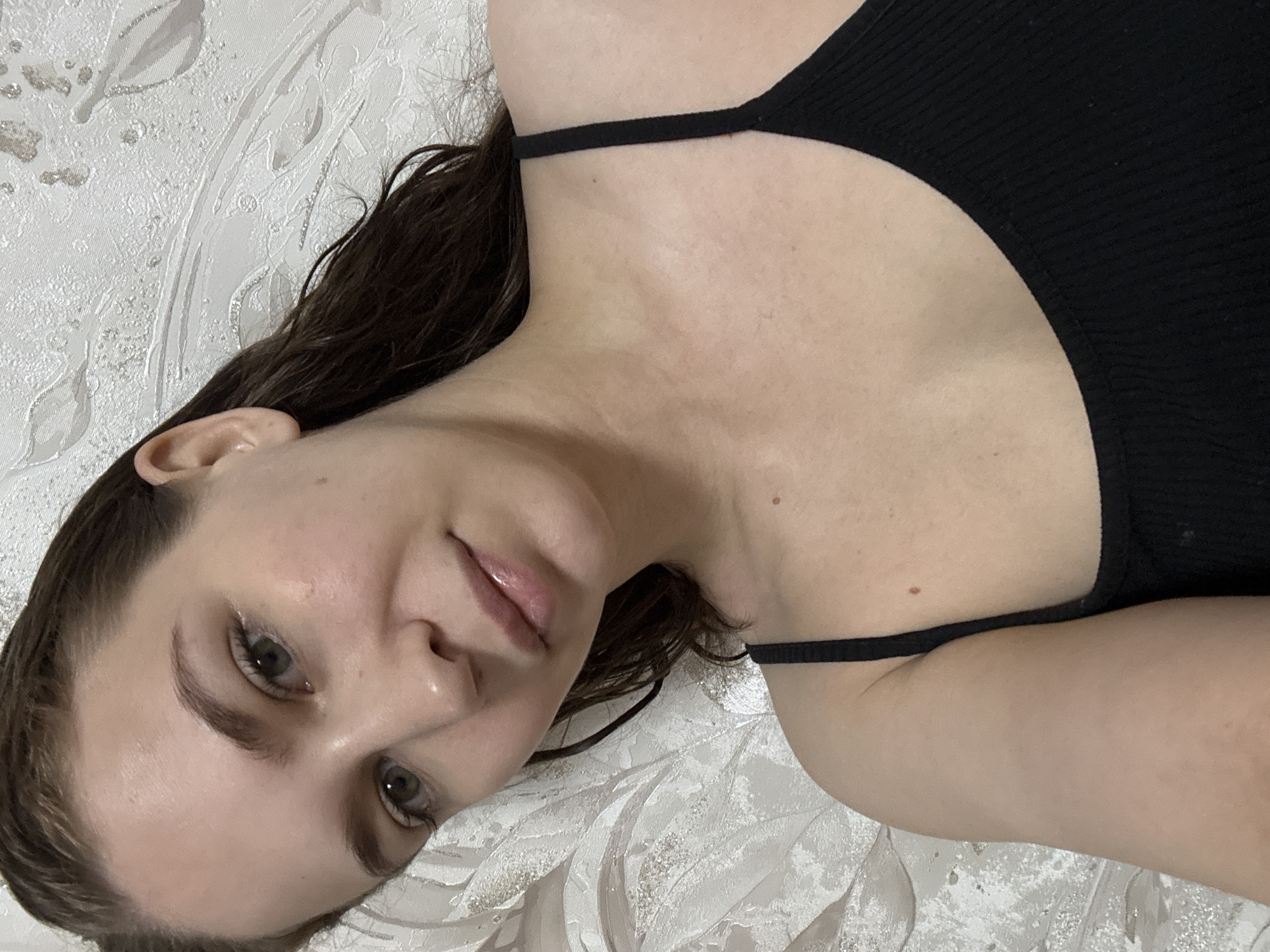 A young woman, appearing to be in her twenties, is depicted in a medium-close, high-angle portrait, taken indoors in Chaikovsky, Russia. She is positioned horizontally, likely lying down, with her head slightly tilted to the left, and her gaze directed towards the camera.
She has long, dark brown hair that appears damp or wet, parted on her right side (viewer's left), with strands falling around her face and shoulder. Her face features light green or hazel eyes, naturally shaped dark eyebrows, a straight nose, and full lips with a subtle gloss. Her complexion is fair with minimal visible imperfections, including a small, light brown mole on her right cheek (viewer's right).
She is wearing a simple black ribbed tank top with thin straps. A distinct, slightly raised brown mole is visible on her left upper chest/shoulder area (viewer's right).
The background consists of a textured, off-white or light gray wallpaper adorned with an embossed floral or leafy pattern featuring subtle silver or metallic accents. The lighting is even and appears to be artificial indoor illumination. No other individuals or significant objects are visible in the frame.