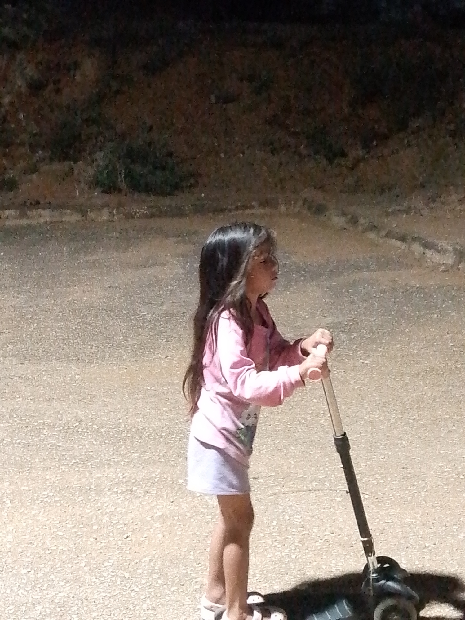 A young girl stands on a scooter outdoors. She is wearing a pink long-sleeve shirt, a white skirt, and white sandals. She holds the scooter handles with both hands. The background is a dark, paved area. The location is likely Juan Griego, Venezuela.