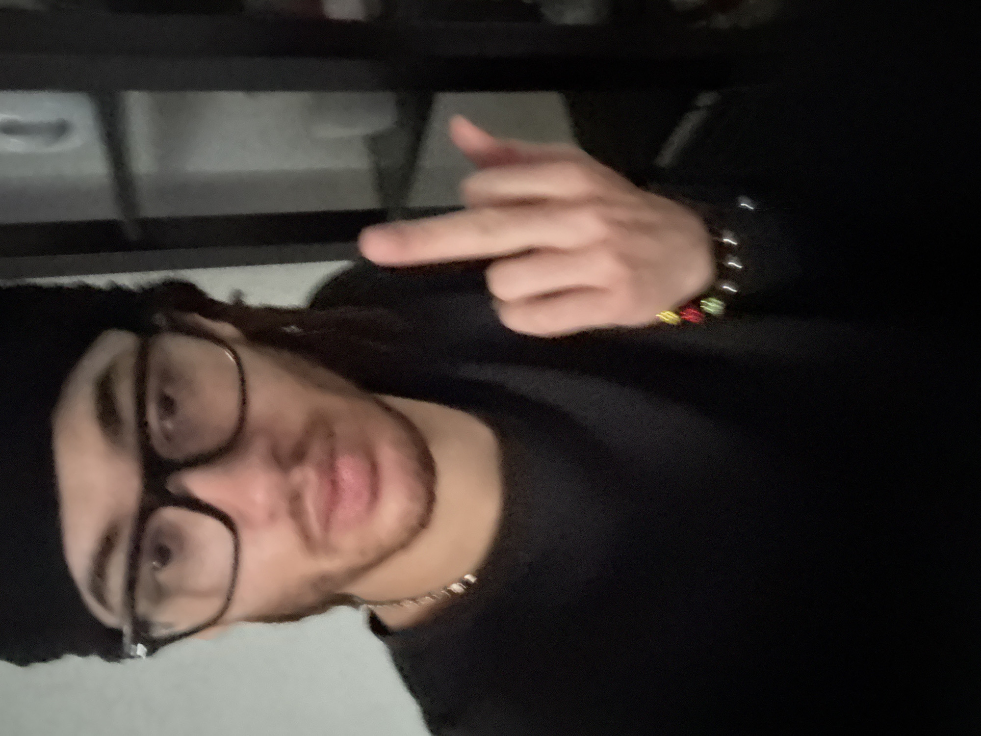 A person with brown skin, wearing glasses and a black hat, and a black shirt is pointing with his right hand. He is also wearing a bracelet on his right wrist and a chain around his neck. The background is blurry and shows some shelves. The location is not discernible from the image.