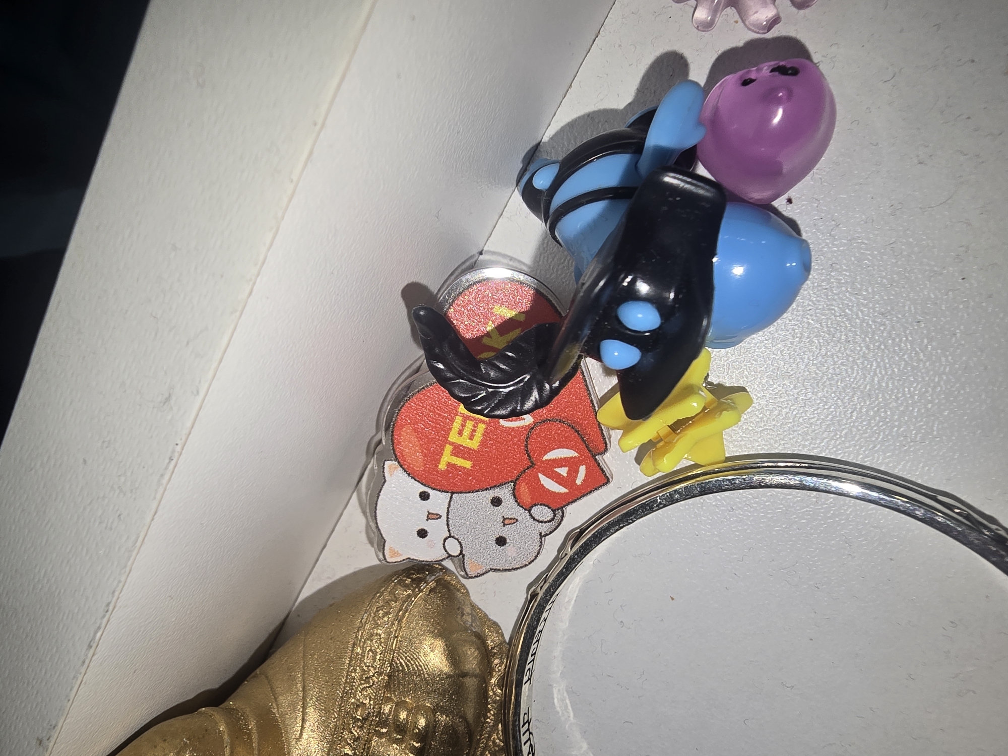 The image shows a collection of small toys and trinkets on a white, textured surface, possibly a shelf. To the left, a portion of a golden, textured object is visible, resembling a bell or a statue. In the center, there's a clear plastic keychain featuring two cartoon cats and a red heart with the letters "TE" and an anarchy symbol. Next to it, a black and blue toy figure with large, round blue eyes and black appendages is present. A small yellow object, possibly a flower petal, is positioned near the blue toy. To the right of the blue toy, a translucent purple object with two small black dots for eyes is visible. The background is blurred, with a dark area above a lighter, textured surface that appears to be the same surface as the foreground. There is no indication of the city of Ulan-Ude, Russia, or any geographical context in the image. The scene is static, with no people or interactions depicted.