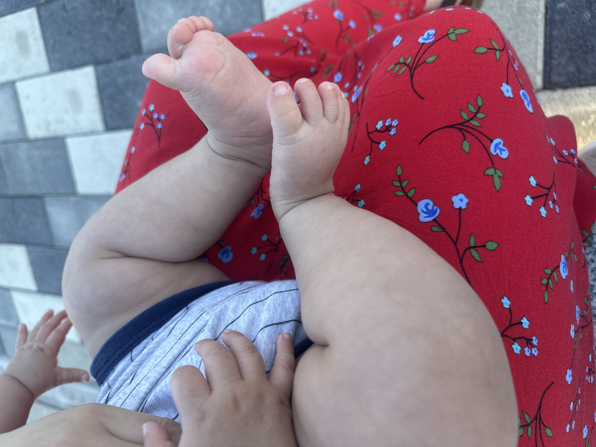 The image shows a close-up of a baby's legs and feet, resting on a red fabric with a blue floral pattern. The baby is wearing light gray shorts with dark blue trim. The baby's feet are small and delicate, with visible toes and fingernails. One foot is positioned above the other, and the baby's legs are bent at the knees. The background consists of a gray and white tiled surface, possibly a patio or walkway. The scene appears to be outdoors, as suggested by the lighting and the texture of the ground. The overall impression is one of tenderness and domesticity. There is no visible text in the image. The location context provided is Manas, Kyrgyzstan, which adds a geographical element to the scene, though it is not directly depicted in the image itself. The time of day and weather are not discernible. The emotion conveyed is likely one of peacefulness or relaxation, as the baby appears to be resting.