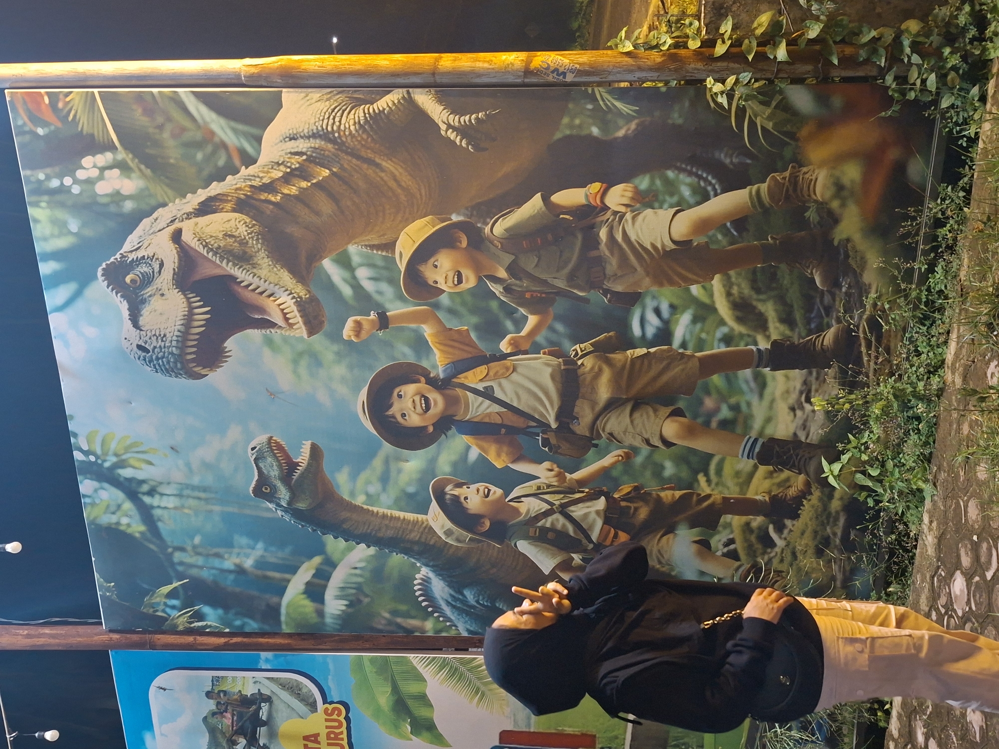 The image shows a person standing in front of a large dinosaur-themed advertisement. The advertisement features three children dressed in khaki explorer outfits, overlaid with dinosaurs, including a Tyrannosaurus Rex and a Brontosaurus. The person in the foreground is wearing a dark hooded jacket and light-colored pants. Some greenery grows behind the advertisement's frame. The picture was taken in Lhokseumawe, Indonesia.