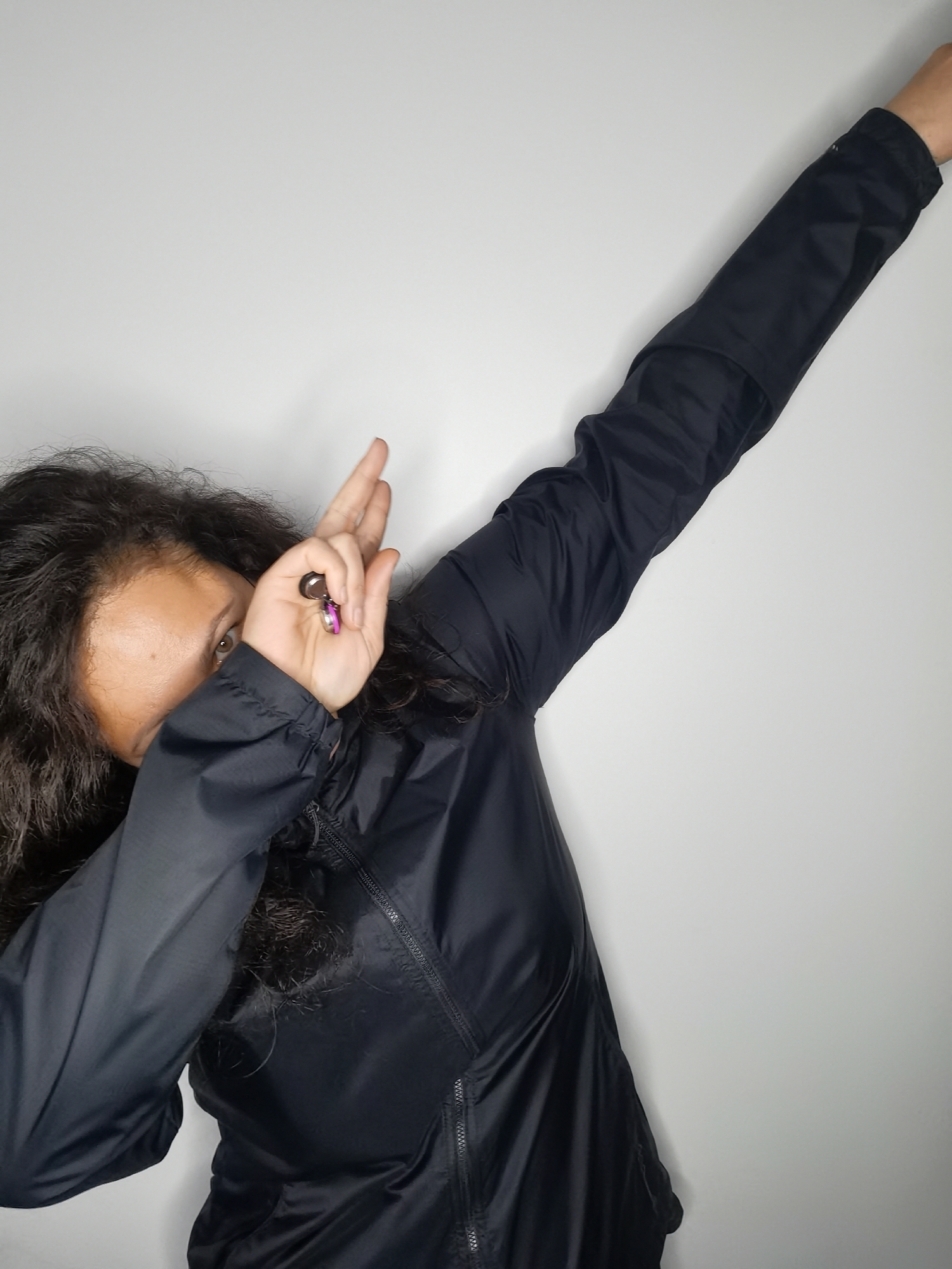 A person with dark, curly hair is shown in a close-up shot, performing a dab pose against a plain white wall. The person is wearing a black jacket with a zipper. Their right arm is extended upwards and to the right, while their left arm is bent at the elbow, covering part of their face. Their left hand is raised, holding what appears to be a keyring with a small charm. The background is a featureless, light-colored wall. The image does not provide any information about the location of Chaikovsky, Russia.