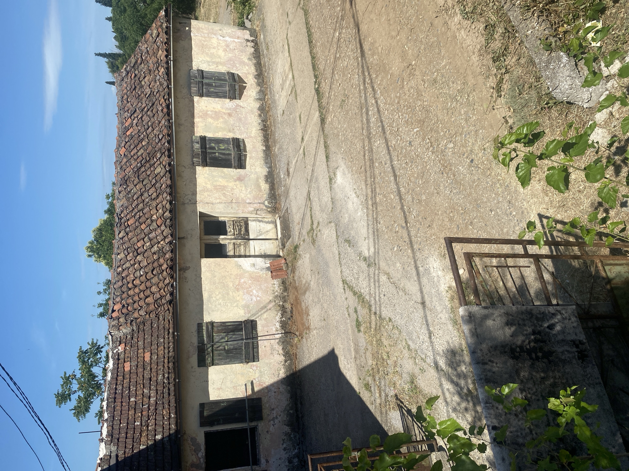 The image shows a weathered, one-story building with a clay tile roof. The building has multiple windows, some with closed shutters. The building’s exterior is aged and shows signs of disrepair. A rough concrete walkway leads away from the building. The ground around the walkway appears to be a mix of dirt and gravel. A rusty metal gate or railing is partially visible in the foreground, partially obscured by greenery. The sky is blue.
Given the architectural style and surroundings, it's plausible this is located in a rural or older part of Podgorica, Montenegro, but without further information, a more precise location cannot be determined.