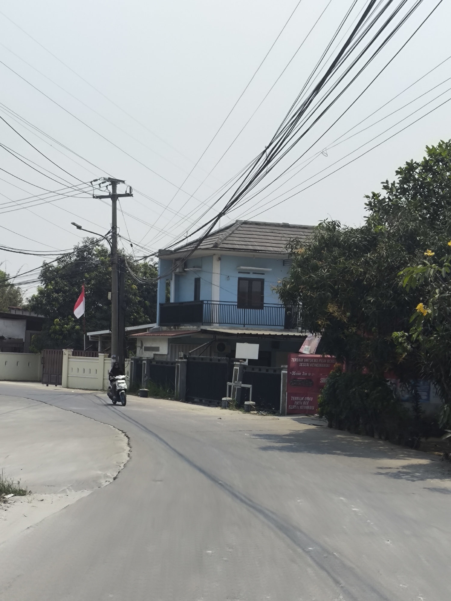 In Cilegon, Indonesia, a moderately paved road curves through a residential area. A person on a white scooter travels along this road, wearing dark clothing and a helmet. To the right, a light blue house with a brown roof and a dark railing on its upper floor stands. A large tree with dark green foliage and yellow flowers is situated in front of the house. The Indonesian flag, red and white, is visible on a flagpole to the left of the house. Overhead, a dense network of electrical wires crosses the sky, supported by a utility pole. The environment appears to be a sunny day with a clear sky.