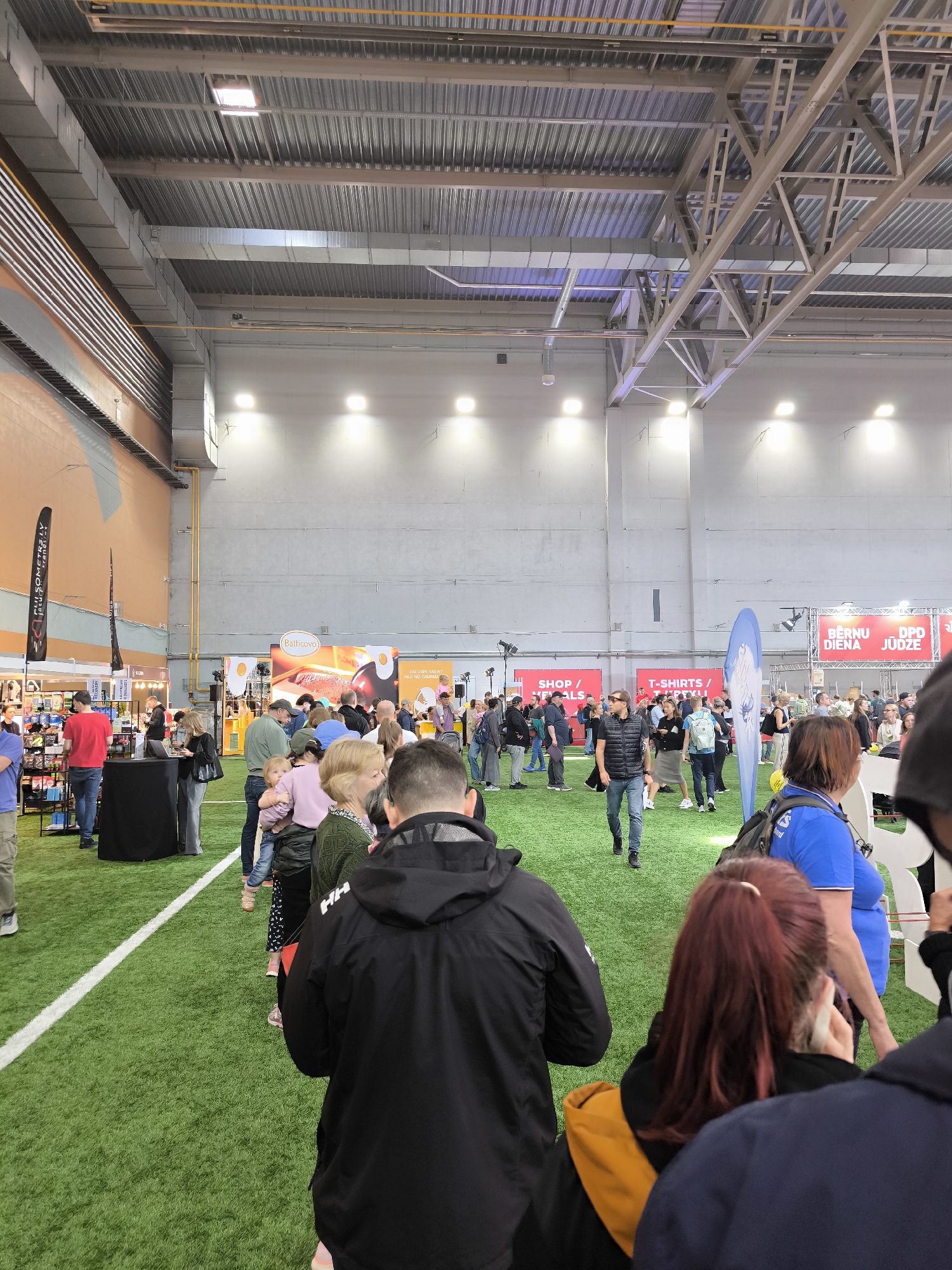 The image shows a crowded indoor event on artificial turf, likely a trade show or exhibition in Riga, Latvia. People are walking and standing near vendor booths with signage in both Latvian and English. Several vendors are visible, offering various products, including food (Balticovo eggs), clothing (T-shirts), and other merchandise. The building has a high ceiling with industrial lighting and metal supports. A line of people extends from one vendor booth, indicating interest in what they are selling or offering. The people are diverse in age and appearance, wearing casual clothing appropriate for an indoor event.