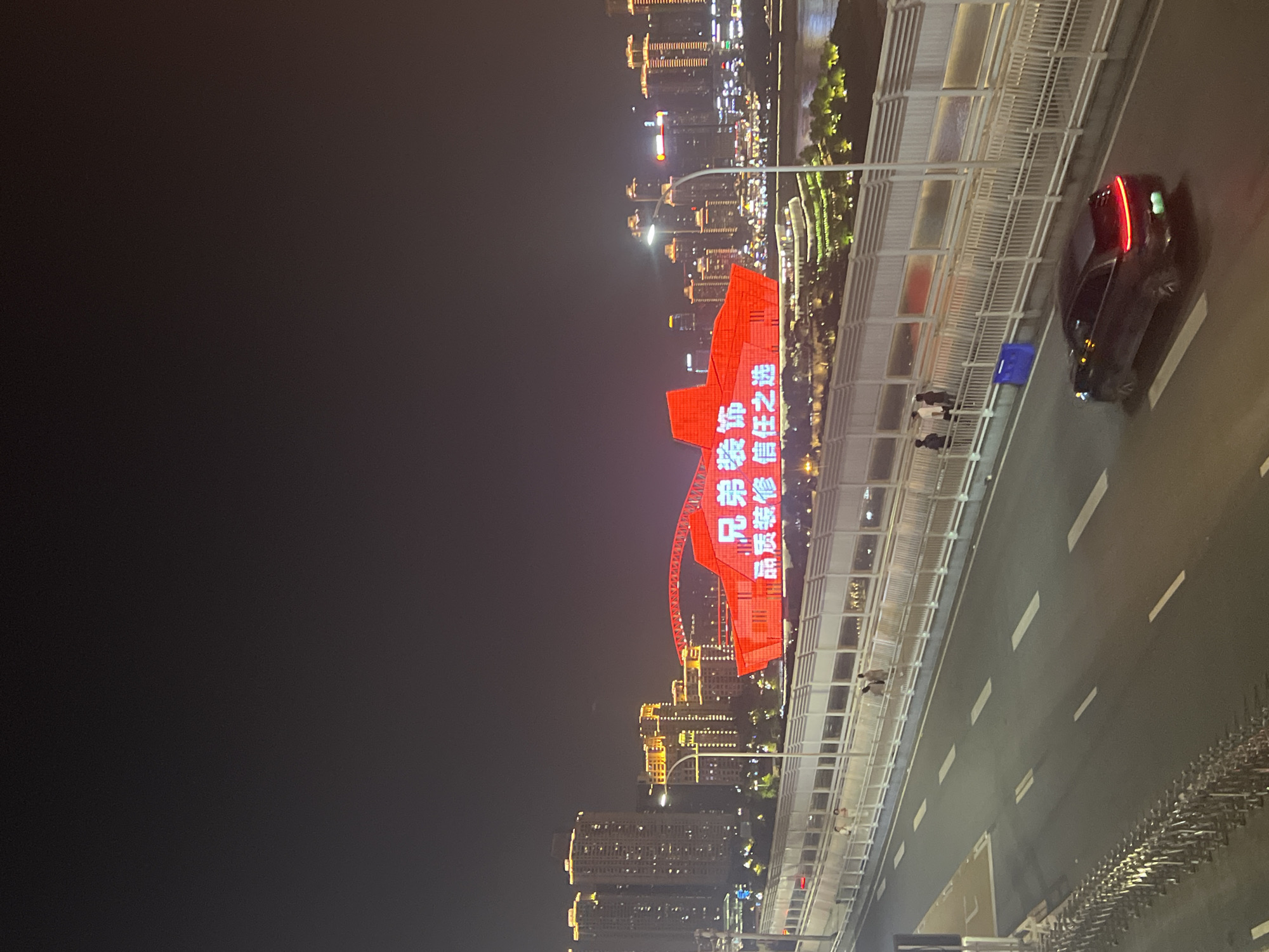 This nighttime, oblique-angle image, taken from an elevated position, depicts a cityscape in Yuzhong District, China. A bridge occupies the foreground, with a black sedan positioned on the right side of the road. Pedestrians are visible on the bridge's walkway. A large red structure with illuminated Chinese characters stands out in the middle ground. The background reveals a city skyline with buildings and lights reflecting off a body of water.