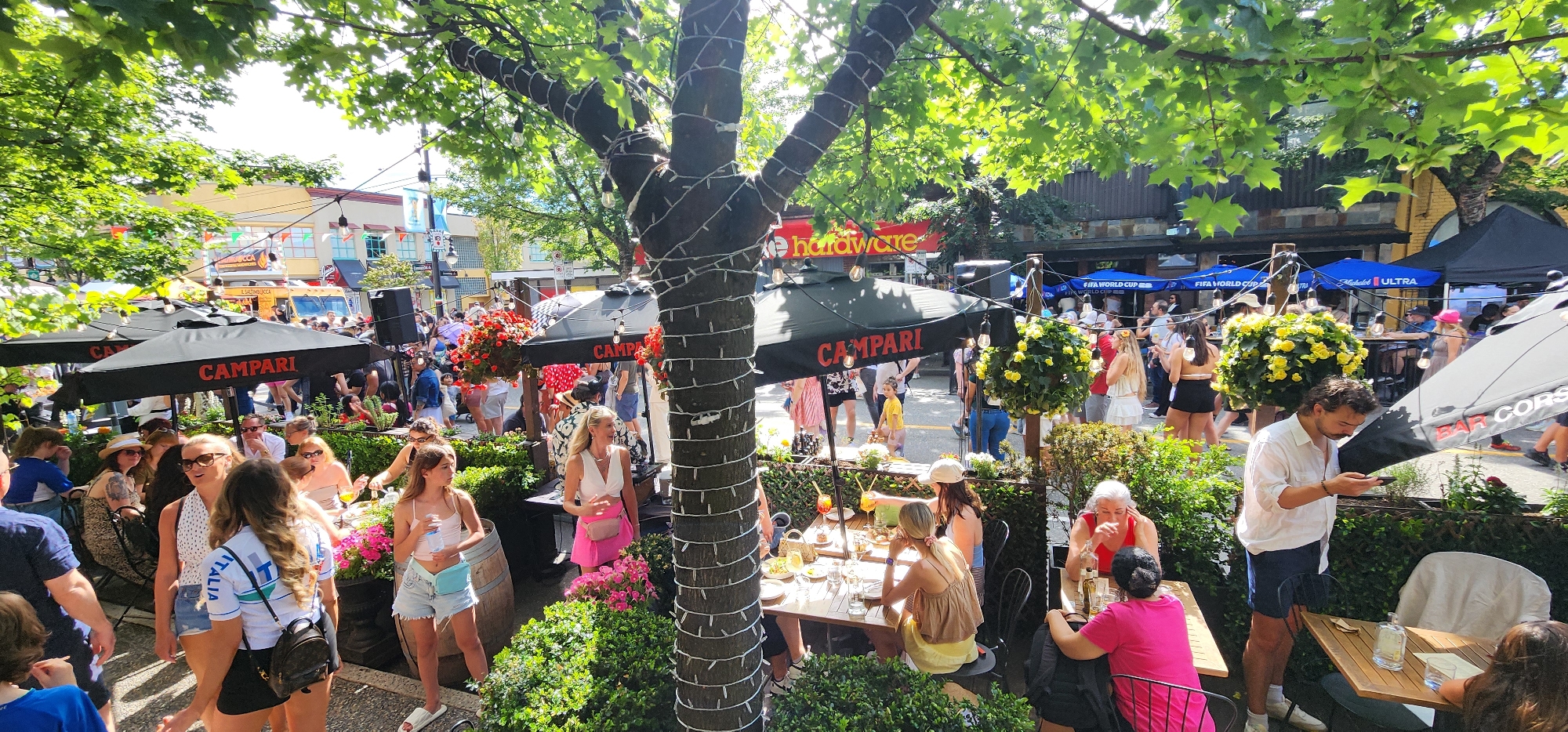 An outdoor patio scene is depicted, filled with people enjoying a sunny day. A tree decorated with string lights stands prominently in the foreground. The patio is furnished with tables and chairs, and several umbrellas are set up, one of which displays the "CAMPARI" logo. In the background, there is a street with buildings, shops, and more people. Food trucks are also visible. Several individuals are seated at tables, engaged in conversation or looking at their phones. Others are standing and milling about. The overall atmosphere appears lively and social, suggesting a public event or a busy restaurant area.