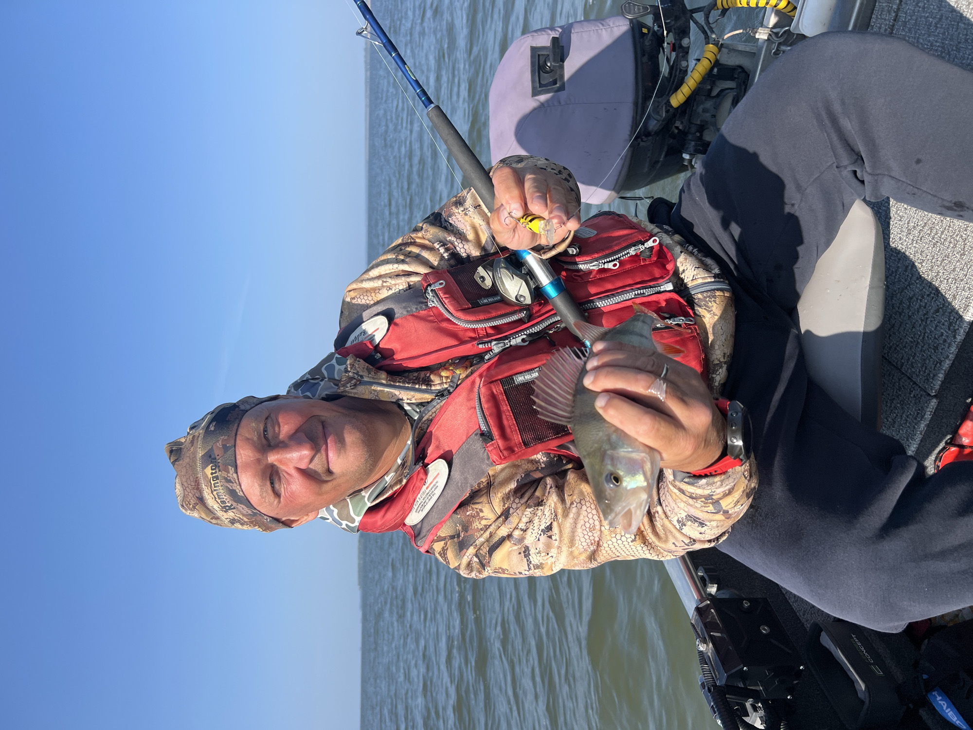 A man is sitting in a boat on a body of water, holding a fish he has caught. He is wearing a camouflage bandana, a camouflage jacket, and a red life vest. He is smiling and looking at the camera. The boat has an outboard motor and fishing rod. The background shows a clear blue sky and a horizon line where the water meets the sky. The location is identified as Borovoe, Russia.