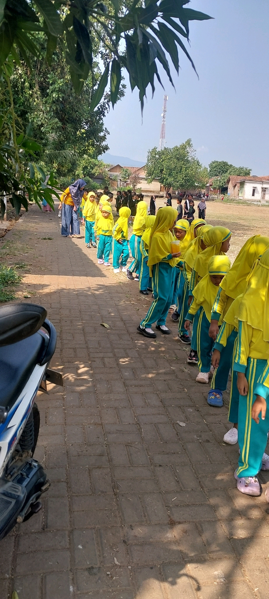 In Keling, Indonesia, a group of children dressed in bright yellow hijabs and teal outfits are standing in a line on a brick-paved path. The children appear to be of elementary school age. Some of them are holding cups. In the background, there are more children, some adults, buildings, trees, and a cell phone tower. A motorcycle is parked on the left side of the path. The overall scene suggests an outdoor activity or school event.