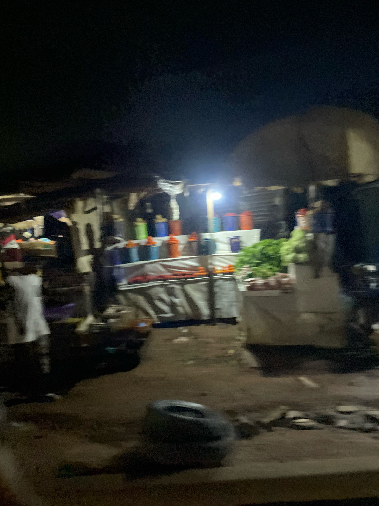 This image depicts a dimly lit outdoor market scene at night in Abuja, Nigeria. The market appears to be a roadside stall with various items for sale, including fresh produce like leafy greens, and an array of colorful buckets or containers, some stacked and others on display. The lighting comes from a bright artificial light source illuminating part of the stall. A person in a white garment is partially visible on the left side of the image. The ground is uneven and appears to be dirt or unpaved road. The overall impression is a bustling nocturnal marketplace, possibly a temporary setup for nighttime commerce.