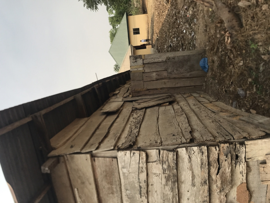 The image depicts an outdoor scene, likely in a rural or semi-urban setting, consistent with Umudike, Nigeria. The foreground is dominated by a weathered wooden structure, possibly a wall or shed, constructed from rough, uneven wooden planks. These planks show significant signs of age, wear, and discoloration, with various shades of grey and exposed grain. The top of this structure appears to have corrugated metal sheeting.
Beyond the wooden structure, a path of dirt and dry ground leads towards a light yellow or tan-colored building in the mid-ground. This building features a sloped, light green corrugated metal roof and a visible rectangular window. Two indistinct figures are standing near this building; one individual is more clearly visible wearing a light-colored shirt. The ground surrounding the pathway and buildings is characterized by dry grass, patches of earth, and scattered debris, including a blue bag or tarp near the wooden structure.
The sky in the background is a uniform, pale grey, suggesting an overcast or hazy day. The lighting is diffused, indicating either midday or late afternoon without direct sunlight. There is no visible text in the image, and no specific activities or emotions are clearly discernible from the figures.
