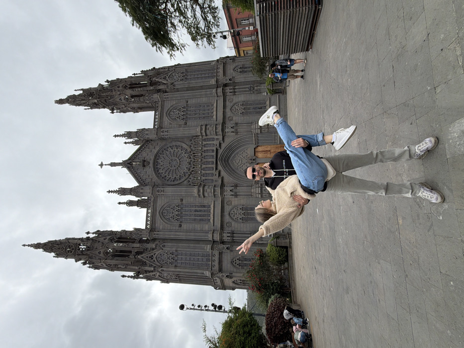 The image captures a daytime scene in a paved public square in Arucas, Gran Canaria, Spain, with the Parroquia de San Juan Bautista (Church of San Juan Bautista) prominent in the background under an overcast sky. In the foreground, a male, estimated to be in his 20s-30s, wearing a black hooded sweatshirt, light grey trousers, and sunglasses, stands holding a female, also estimated to be in her 20s-30s, horizontally in his arms. The female, dressed in a beige fluffy sweater and light blue jeans, has her right arm raised in a peace sign gesture and is looking at the male. The male is looking at the female. The church behind them is a dark grey neo-Gothic structure with two prominent spires, a large central rose window, and multiple ornate arches and windows. To the far left, a group of individuals, including children, are visible near green bushes and a multi-headed lamppost. To the right, two adult individuals stand in the mid-ground, with a stack of wooden pallets and a building with red and white facades further in the background.