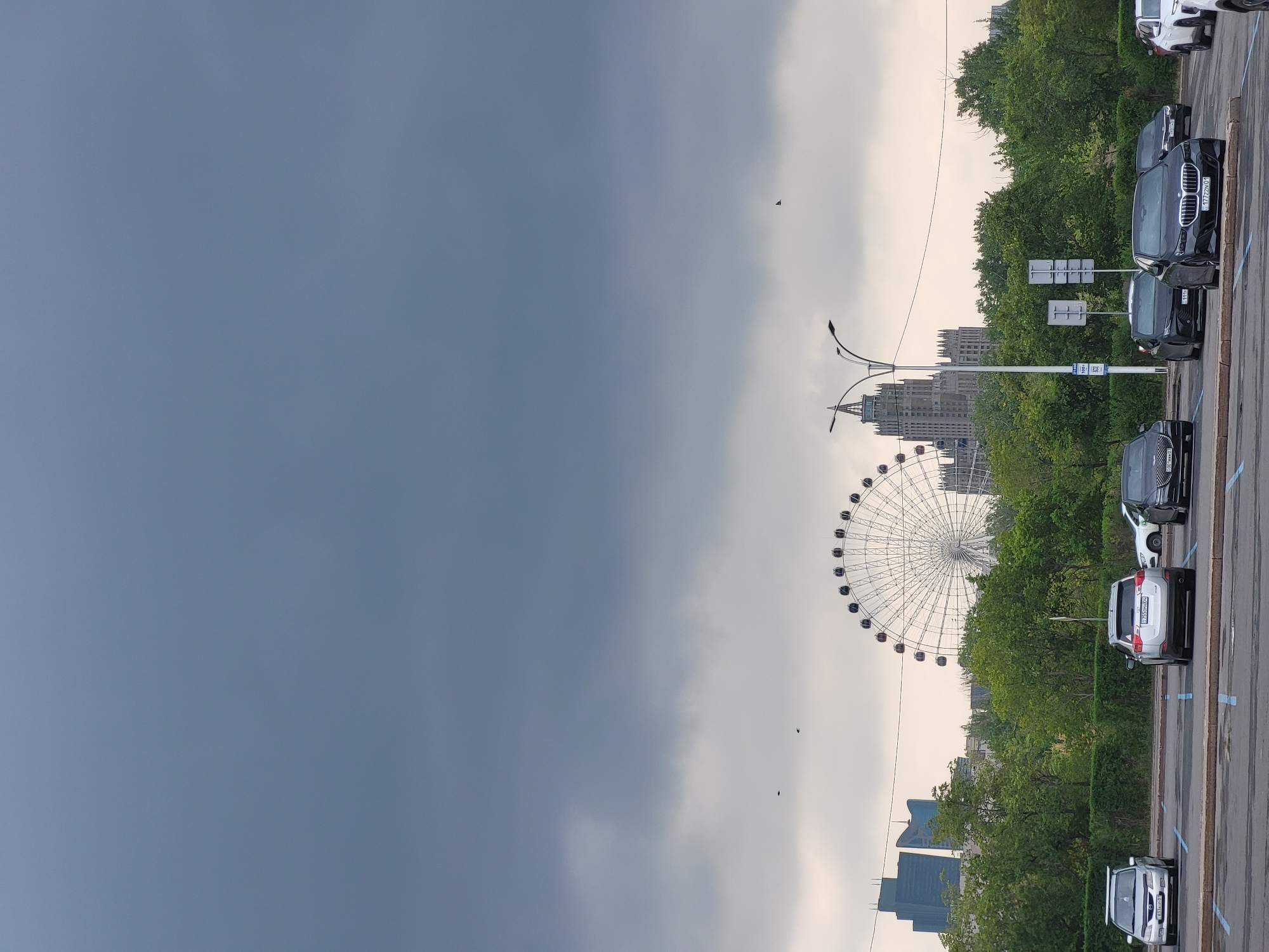 The image depicts a cloudy day in Astana, Kazakhstan. In the foreground, several cars are parked in a lot. The cars include a silver minivan, a silver SUV, and several dark-colored sedans. In the middle ground, there is a line of trees and hedges, with two modern skyscrapers visible in the distance behind them. A large Ferris wheel stands prominently to the right of the skyscrapers. To the right of the Ferris wheel, a tall, gothic-style building with a spire is visible. Several streetlights are also present in the middle ground. The sky is overcast with dark clouds.