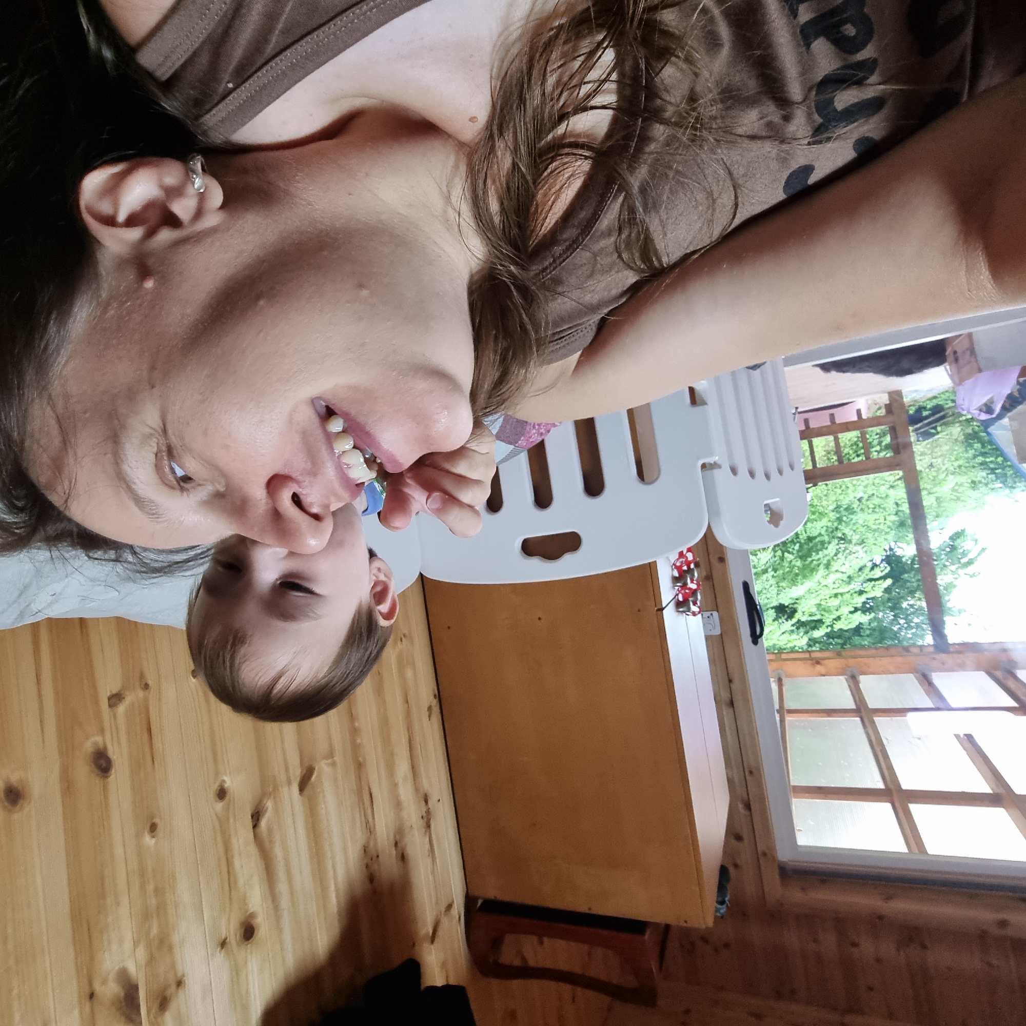 The image shows an overhead, rotated view of two people, an adult and a young child, lying down on a wooden floor. The adult, who appears to be female, is on the left side of the frame, with her head near the top. She is wearing a brown t-shirt. The child is positioned beneath the adult, with their head closer to the bottom of the frame. The adult is looking towards the child and smiling, with her mouth open, revealing teeth. The child is also looking towards the adult.
Behind the people, there is a white plastic playpen. Further in the background, there is a wooden cabinet and a window with a view of green trees and foliage. The scene appears to be indoors, with the wooden floor and the interior elements suggesting a home or a cabin-like setting. The lighting suggests it might be daytime, possibly late morning or afternoon, given the natural light coming through the window. The overall mood seems relaxed and playful. There is no visible text in the image.
