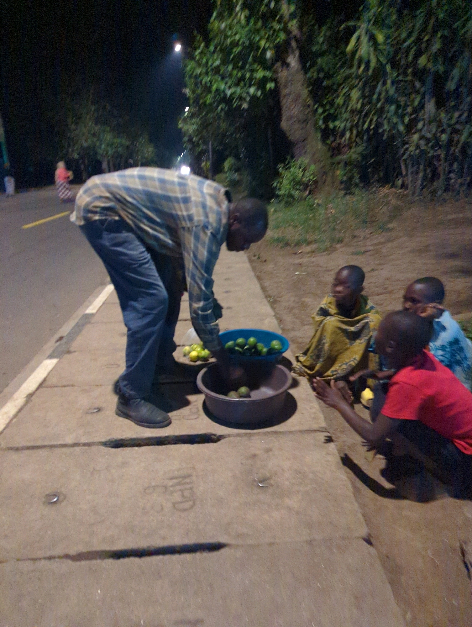 An adult male, wearing a plaid shirt and blue jeans, is bent over a collection of green fruits in two containers on a concrete sidewalk at night. He appears to be selling the fruit to three children who are seated on the ground nearby. One child is wearing a yellow patterned garment, another a light blue shirt, and the third a red shirt. The sidewalk is adjacent to a paved road with a yellow line. Trees and foliage are visible in the background, illuminated by artificial lighting. The scene takes place in the Musanze District of Rwanda.
