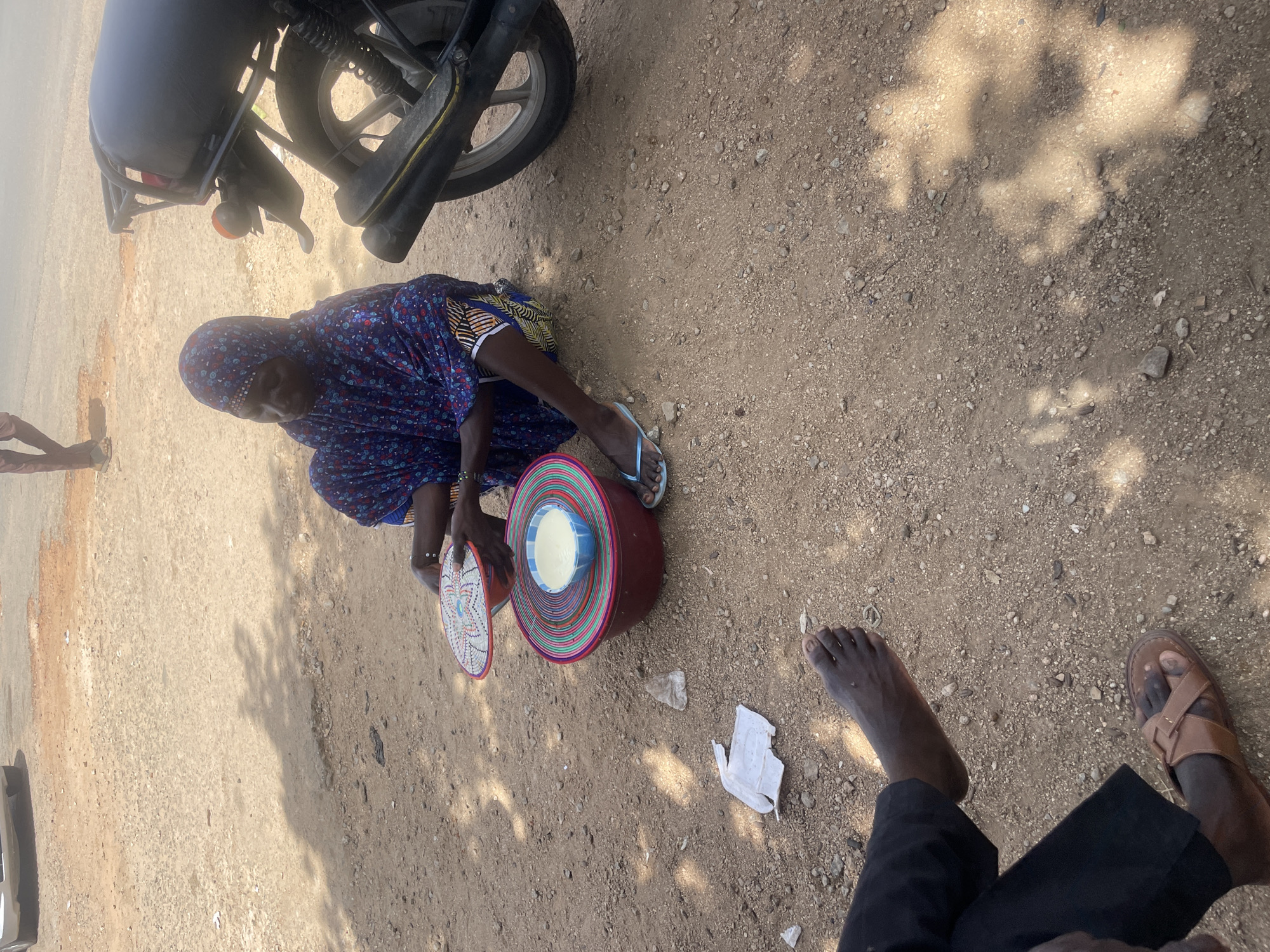 A person wearing a blue headscarf and a patterned dress is sitting on the ground next to a motorcycle. They are holding a woven circular object in their hands and appear to be interacting with a large, round, maroon container that is filled with a pale yellow substance. The container has colorful striped bands around its exterior. The ground is covered with dirt and small stones, and there are shadows from trees cast across the scene, suggesting it is daytime. Another person's feet, one bare and the other wearing a brown sandal, are visible in the lower right corner of the image. A piece of white paper is lying on the ground. The overall scene suggests a casual outdoor setting, possibly a roadside market or resting spot. There is no visible text in the image.