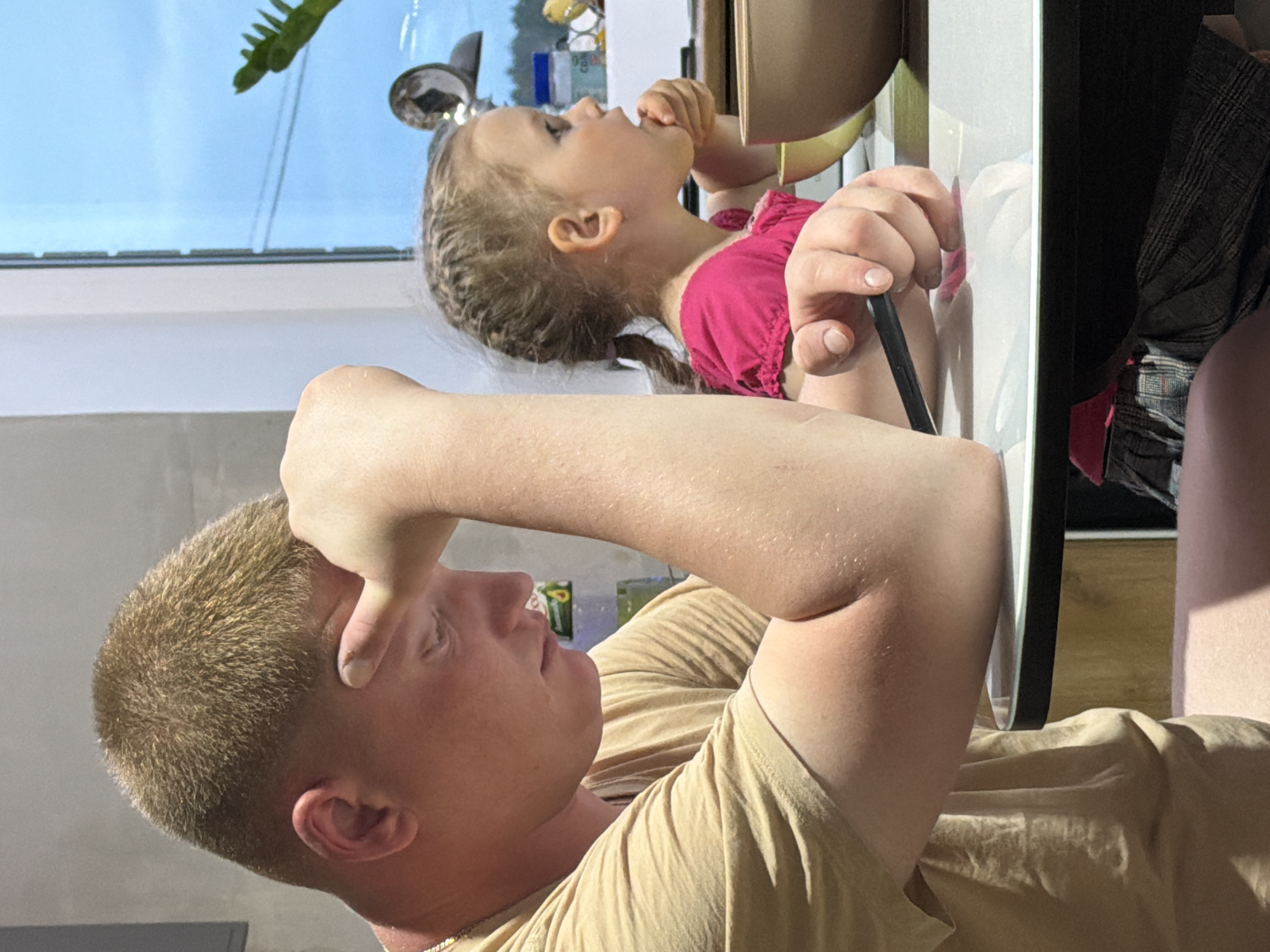 A man and a young girl are at a table. The girl, in a pink shirt, is eating with her hand, and the man, in a tan shirt, is shielding his eyes with his hand. There is a window to the left with a plant, and various objects are on the window sill. The image was taken indoors. The exact location in Chaikovsky, Russia, cannot be determined from the image.