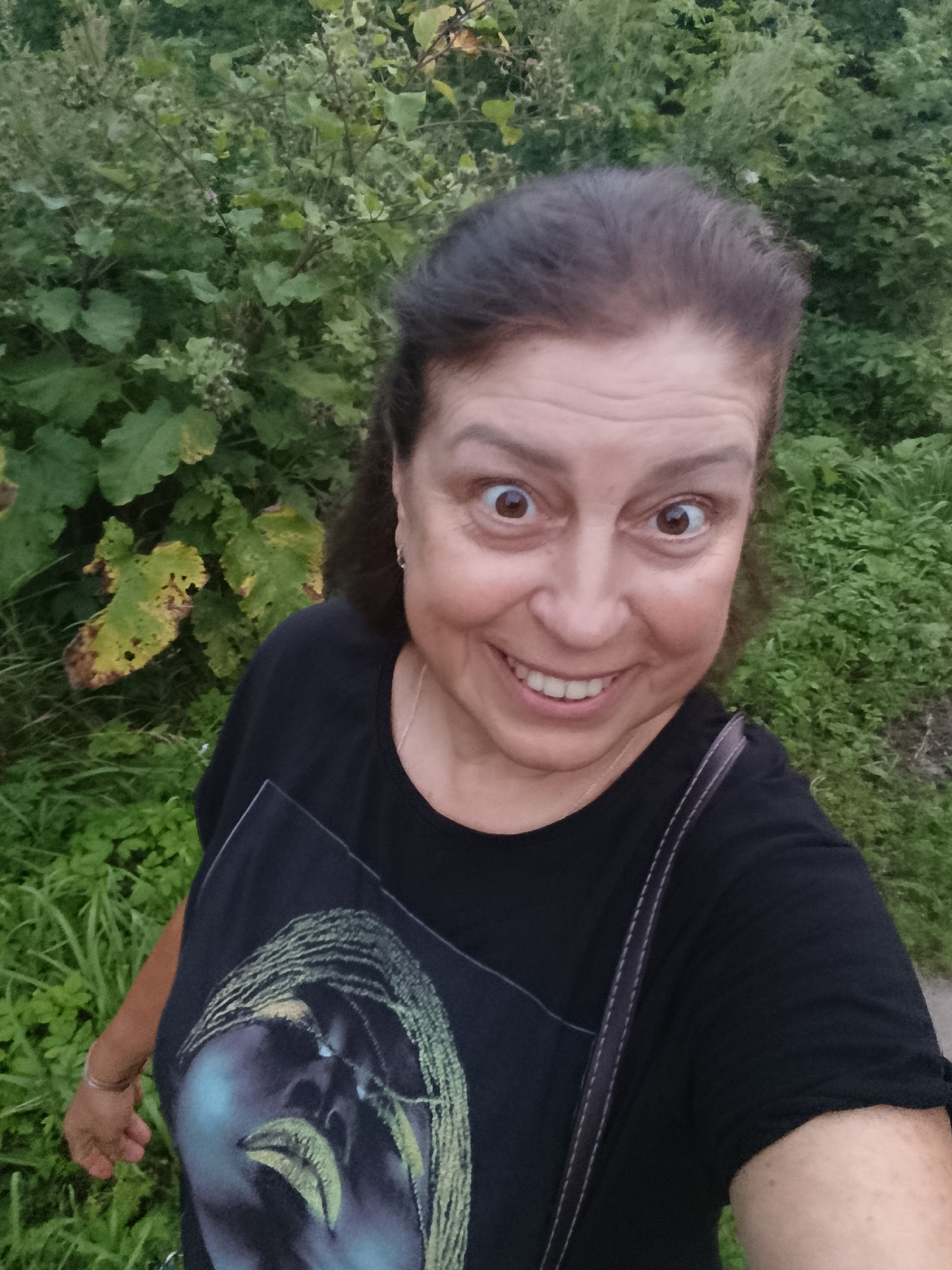 A medium shot captures a smiling woman from the chest up, taken outdoors. She has dark hair pulled back and is wearing a black t-shirt with a graphic of a face. The background consists of lush green foliage, suggesting a natural, possibly wooded, setting. The woman's eyes are wide and her mouth is open in a broad smile, with her teeth showing. Her skin appears fair, and she is wearing a thin silver bracelet on her left wrist. A dark strap, likely from a bag, is visible across her shoulder. The lighting appears natural and somewhat dim, indicating it might be evening or an overcast day. The image does not contain any specific landmarks or elements that would allow for precise identification of the location within the city of Vereya, Russia, beyond the general context of an outdoor environment.