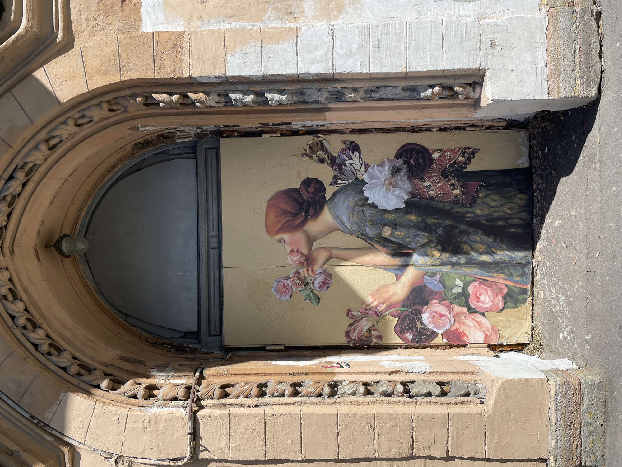 This media file captures a close-up view of a mural on the exterior of a building, situated in Saint Petersburg, Russia. The mural depicts a woman with red hair, wearing a dark, patterned garment, smelling a bouquet of pink and purple flowers. She is positioned within an arched architectural feature made of light brown stone. The stone work surrounding the mural is ornate, with carved leaf motifs. The mural itself is applied to a light yellow surface.
The mural appears to be composed of various cutouts, giving it a collage-like appearance, with a white flower, a pomegranate, and a patterned fabric element also incorporated into the composition. The building's facade shows signs of age and wear, with chipped paint and some sections of plastering visible. The sunlight suggests it is daytime. The overall impression is one of artistic decoration integrated into an old urban environment.