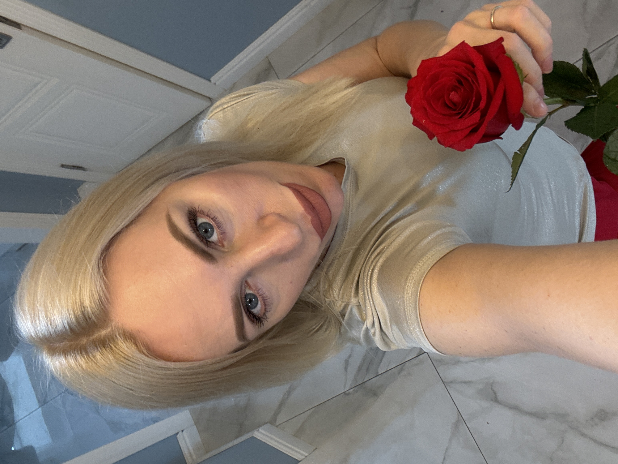 The media file is an indoor, overhead selfie-style photograph of a young woman. Her blonde hair is parted on the side and frames her face. She has light blue eyes and is looking directly at the camera with a calm expression, her lips slightly parted. She is wearing a light-colored, possibly shimmery or metallic, short-sleeved top. Her right hand, adorned with a silver ring on the ring finger, holds a single vibrant red rose by its stem. The background shows a light-colored, possibly white, paneled door in the upper left, and a blue wall segment. The floor is covered with light grey and white marbled tiles. The overall lighting is even, suggesting an indoor setting. There is no visible text.