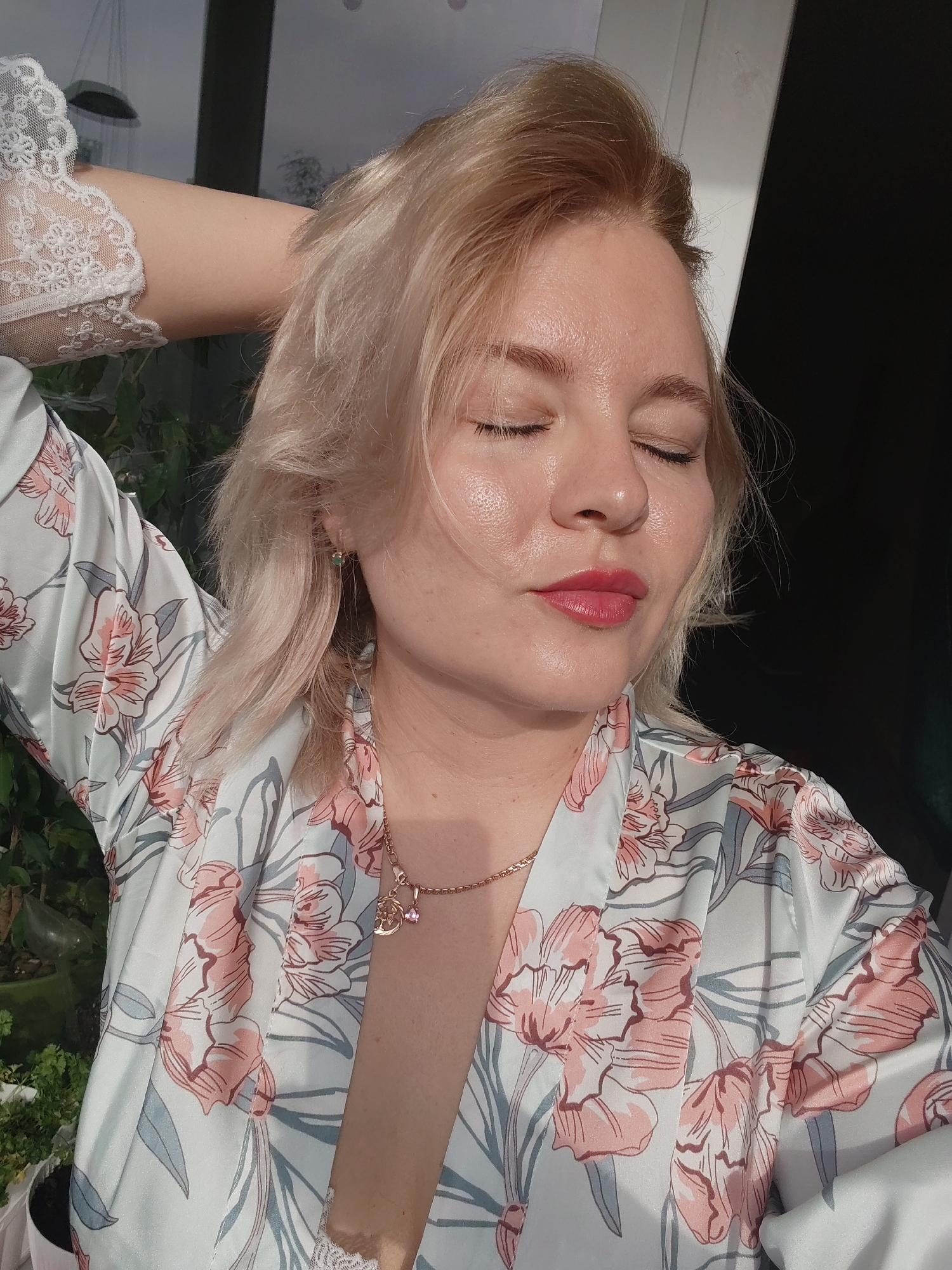 A fair-skinned woman with short blonde hair is centered in the image, eyes closed. She is wearing a light-colored robe with a pink and gray floral pattern, revealing a white lace undergarment beneath. A gold necklace adorns her neck. Her left arm is raised, hand behind her head.
The background shows a blurred mix of greenery and a structure or window frame that is light in color on the left and darker on the right. There is no indication of this scene taking place in Izhevsk, Russia.