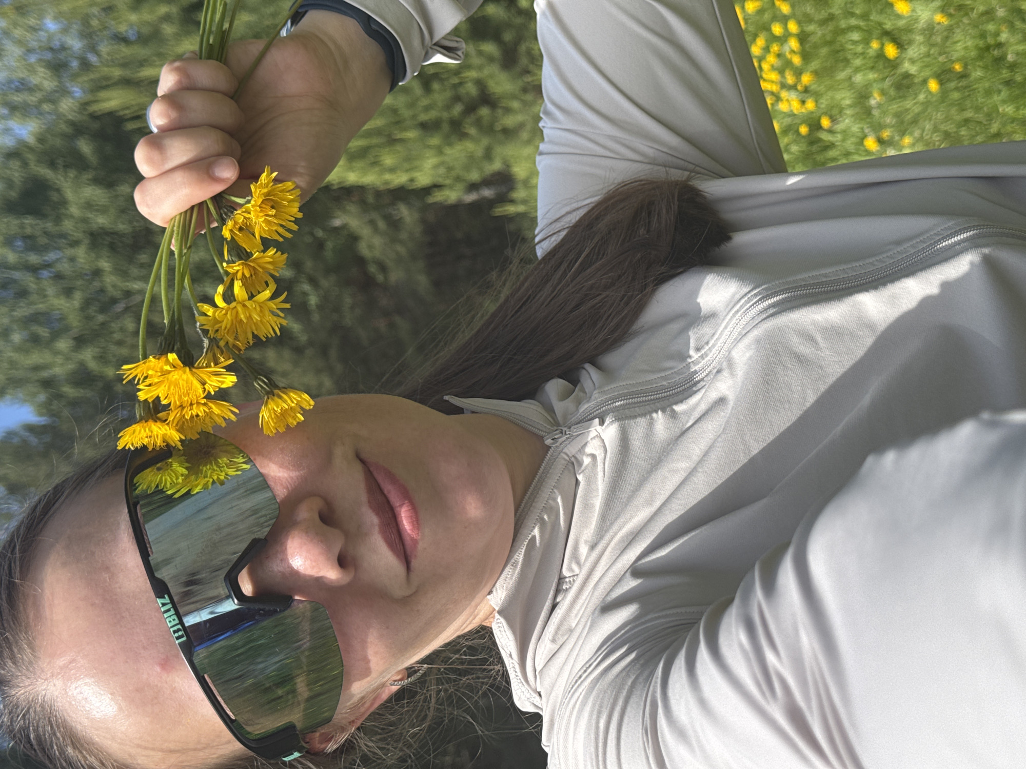 The image captures a person, likely female, relaxing outdoors on a sunny day in what appears to be a dandelion-filled field in Chaikovsky, Russia. The shot is taken from a low angle, with the person lying down.
The main subject is a woman with long, dark brown hair and fair skin, displaying a subtle, contented smile. She is wearing large, reflective, sporty sunglasses, with the brand name "BLIZ" visible on the arm. Reflected in her sunglasses are the green grass, yellow dandelions, and possibly a path or road, along with the bright blue sky.
She is dressed in light-colored (appearing beige or light grey) casual attire, consisting of a zipped long-sleeved top or jacket and matching pants. A watch or fitness tracker is visible on her left wrist. Her right hand, raised, holds a fresh bouquet of bright yellow dandelions.
The setting is an expansive outdoor area, characterized by lush green grass carpeted with numerous yellow dandelions, suggesting springtime or early summer. Trees are visible in the background, under a clear blue sky. The presence of strong sunlight indicates it is daytime, casting visible shadows. The overall scene conveys a feeling of relaxation and appreciation for nature.