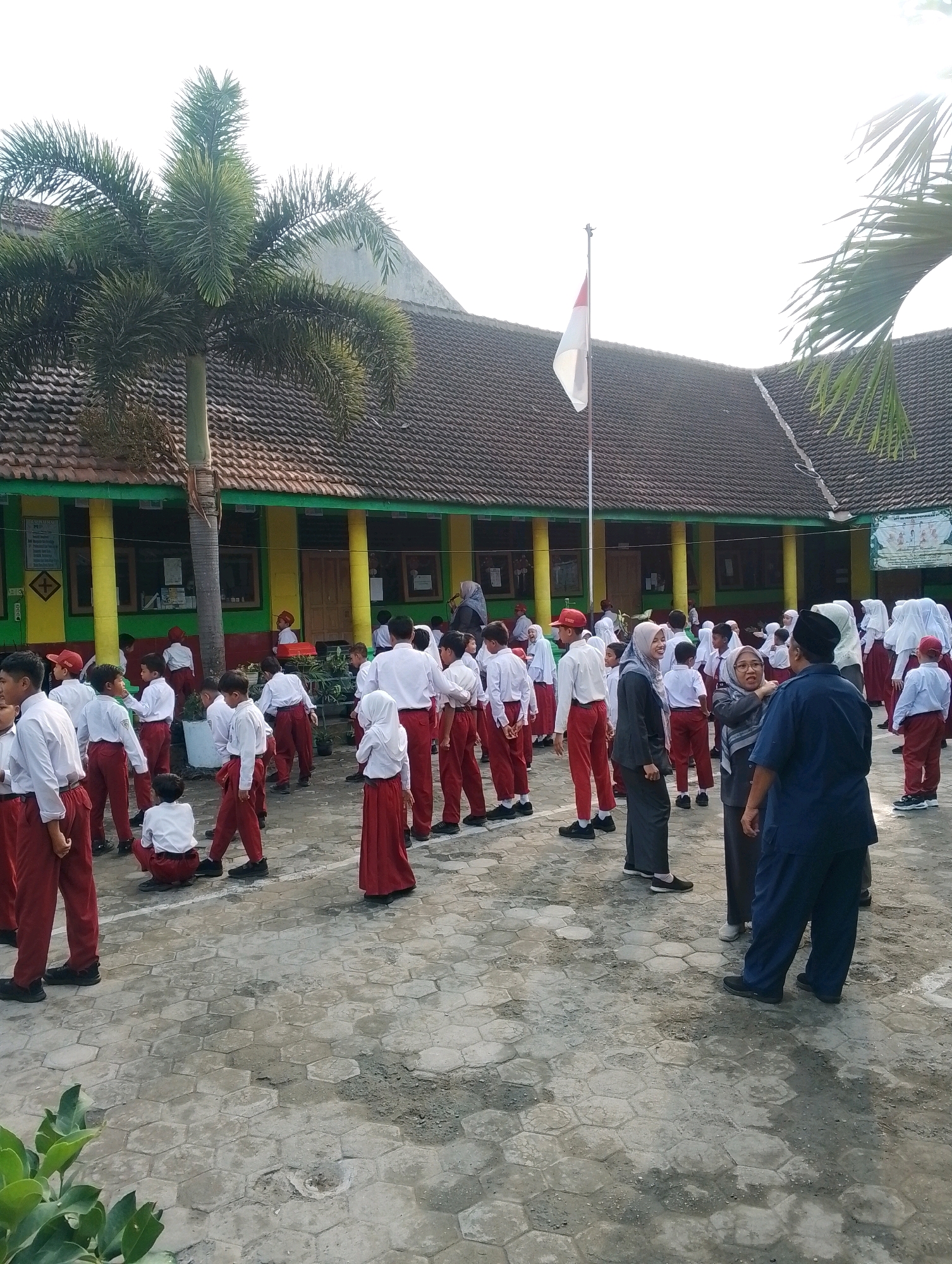 In Papungan, Indonesia, at a school, a group of young students in white shirts and red pants are gathered outdoors. Some students are facing away from the camera, while others are interacting with each other. A few adults are also present, including a woman in a headscarf and a dark blue uniform, and another woman in a grey suit and headscarf, who appears to be in conversation with a man in a dark blue uniform. In the background, a building with a tiled roof and yellow and green accents is visible, along with a palm tree and a flag pole flying the Indonesian flag. The ground is paved with hexagonal tiles. The overall scene suggests a school gathering or assembly taking place in an outdoor courtyard.