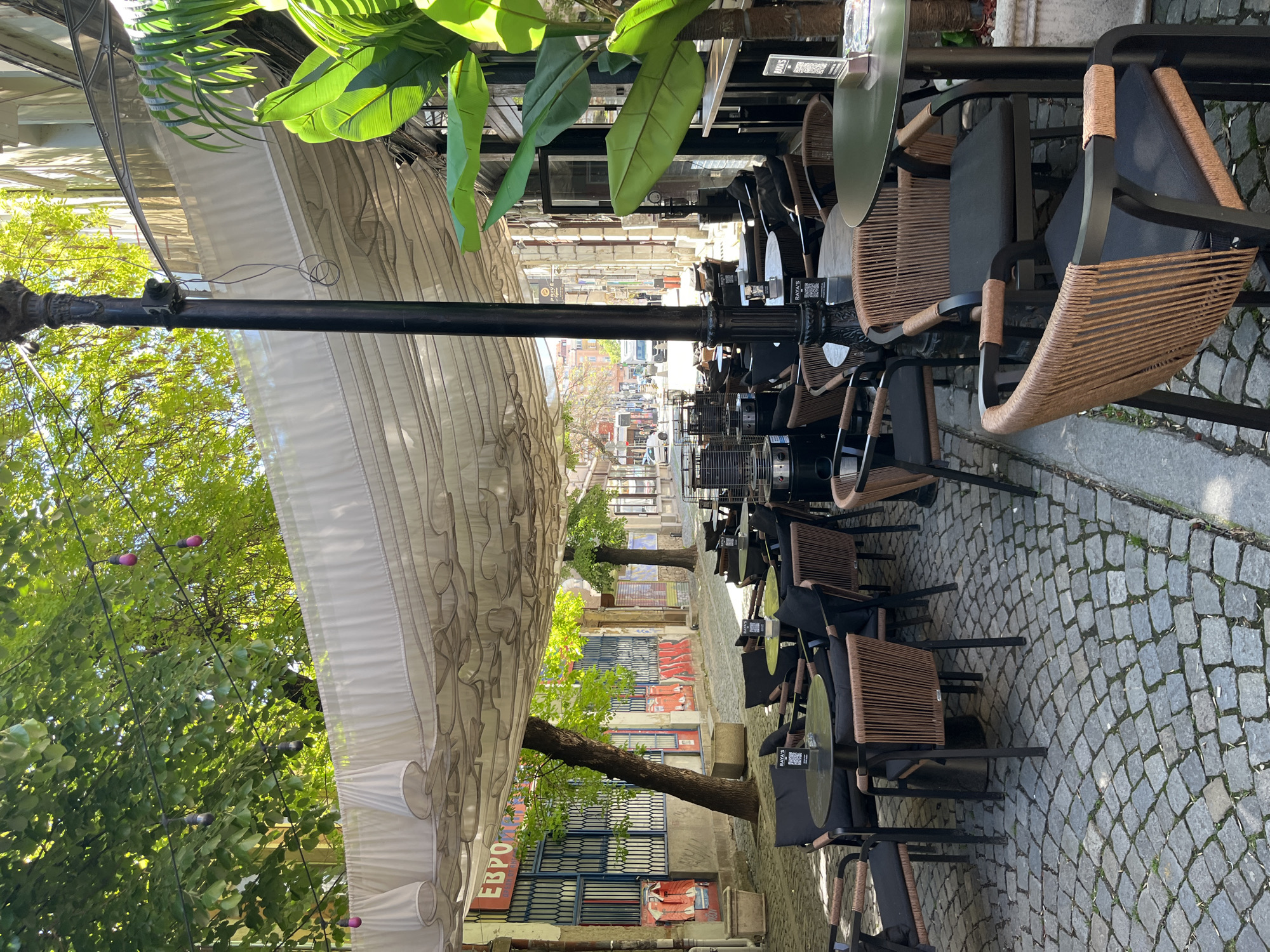 The image depicts a street scene in Plovdiv, Bulgaria, likely a pedestrian area. The foreground is dominated by a row of outdoor cafe tables and chairs, set on a cobblestone street. The chairs are mostly dark metal with woven brown seats. The tables are small and appear to be dark-colored metal with some having QR codes. A retractable awning covers the tables, supported by a black metal pole visible in the center of the image. The background shows a street lined with buildings of varying heights and architectural styles, some with signs and advertisements in Bulgarian. Trees and greenery are visible on both sides of the street, adding to the ambiance. The scene is bathed in daylight, suggesting it was taken during the day. There is no visible human presence in this image.