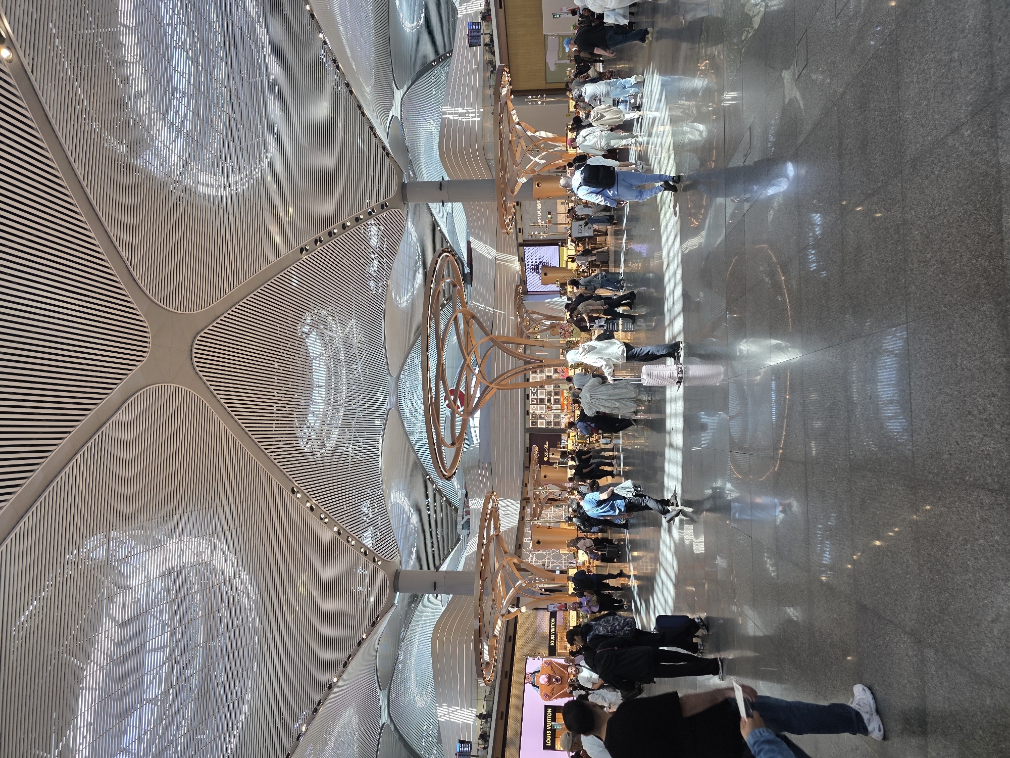 The image captures an interior scene at Istanbul Airport, located in Arnavutköy, Turkey. The perspective is tilted, emphasizing the architectural details of the ceiling. The ceiling is constructed with a white, linear pattern, creating a latticework effect. People are seen walking along a brightly lit corridor lined with shops and stores. Individuals are focused on their own tasks, such as walking and shopping. A circular, decorative structure is a focal point, positioned in the center. The floor is a polished, light-colored material that reflects the ceiling’s design and the surrounding light.