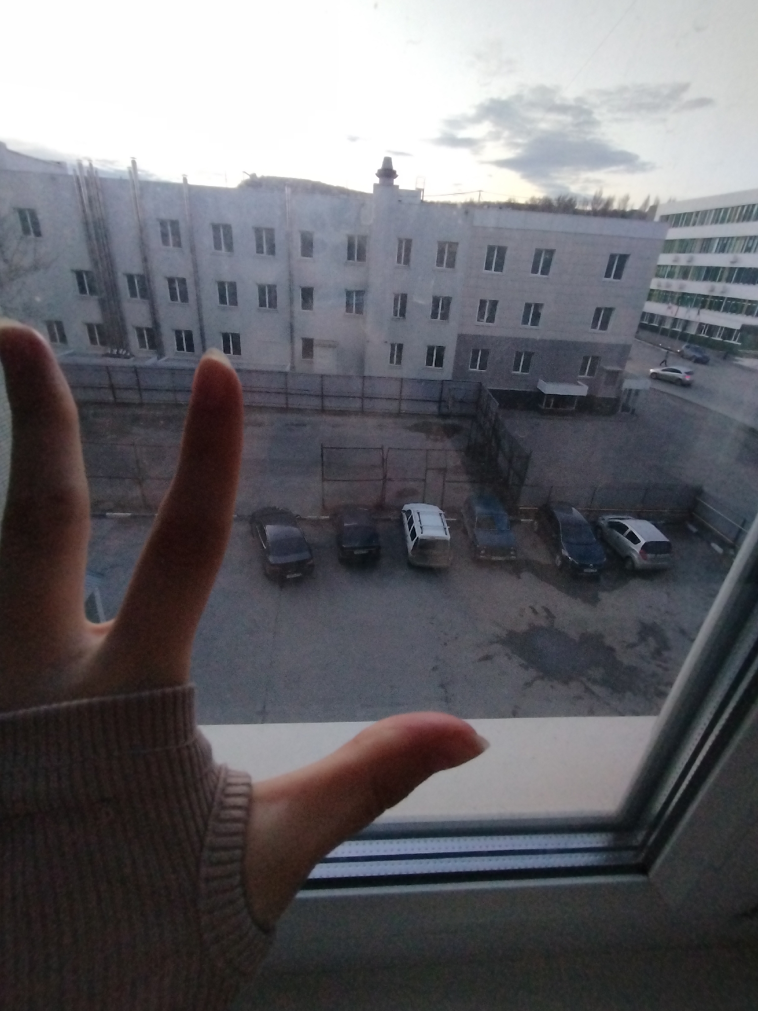 The image shows a view from a window, likely in Saratov, Russia based on the prompt. Outside the window is a parking lot with several cars parked. Behind the parking lot is a large, multi-story building with many windows. To the right of the building is another building with a modern architectural design. The sky is overcast. In the foreground, there is a hand with the index and middle fingers extended, making a peace sign. The hand is partially covered by a beige-colored sweater sleeve. The window frame is visible on the right side of the image.