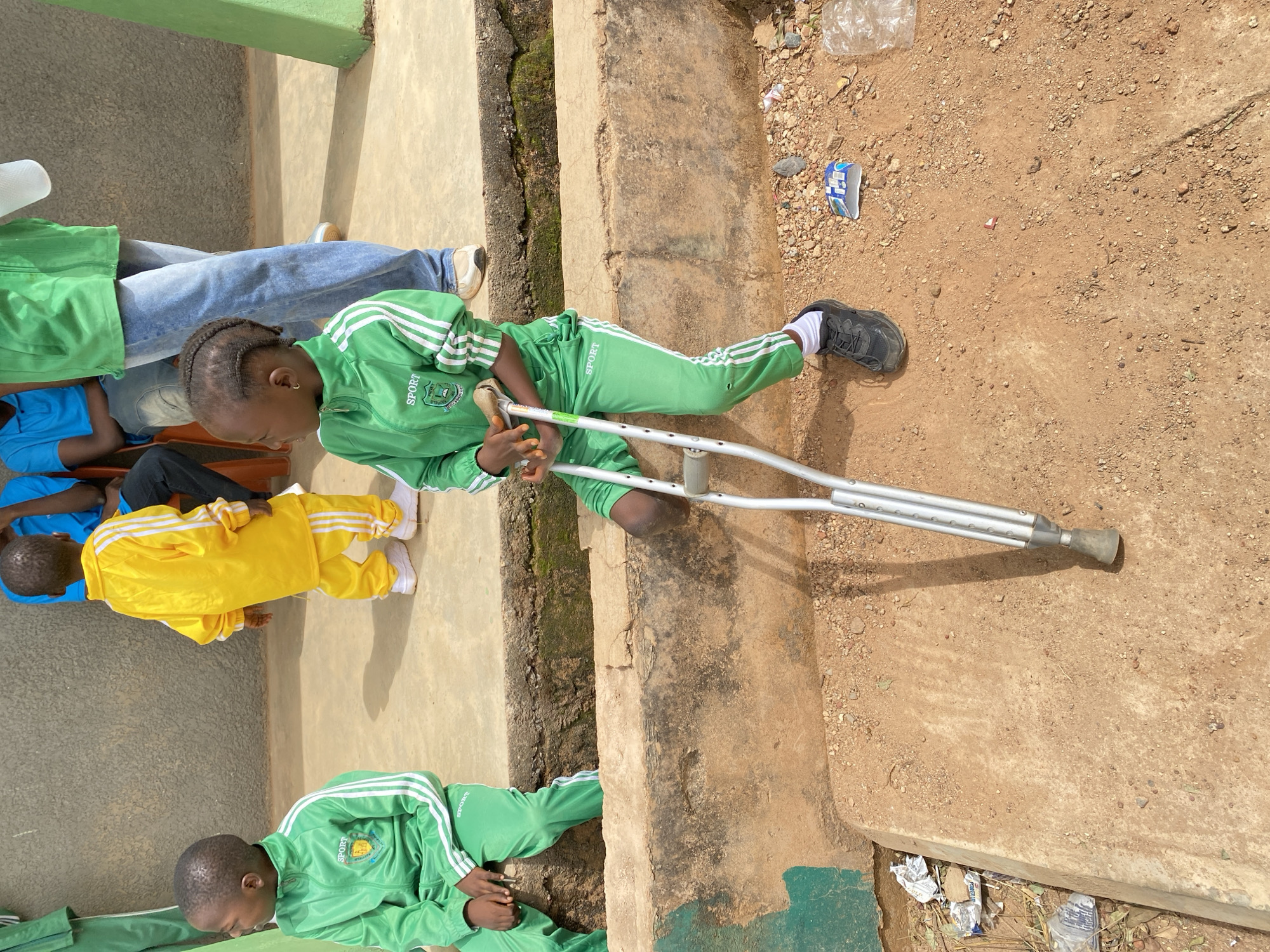 The image, taken in Gura Top, Nigeria, shows a group of children sitting on or near a concrete step. One child in a green tracksuit sits with one leg extended and a crutch, suggesting a possible leg injury. Other children are wearing blue and yellow tracksuits. The ground adjacent to the step is dirt with some debris. A small amount of garbage is visible nearby.