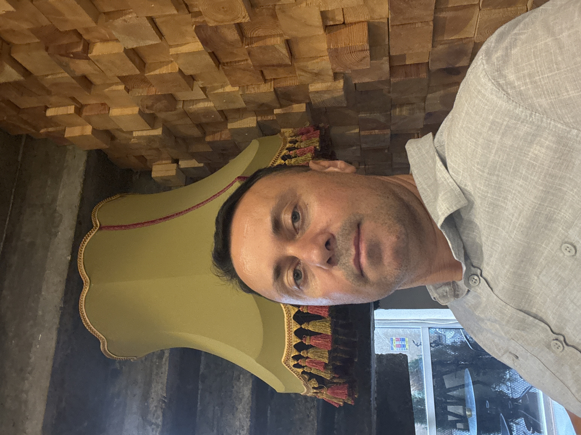 A medium shot selfie is taken from chest level, featuring a man with short, dark hair and a light grey collared shirt. He is positioned in the foreground, with his face occupying the upper portion of the image. Behind him, a distinctive yellow lampshade with red and yellow tassels hangs from the ceiling. The background is dominated by a wall covered in a mosaic of wooden blocks, arranged in a staggered pattern. To the left of the lampshade, a window offers a glimpse of an outdoor seating area with tables and chairs, suggestive of a cafe or restaurant. The architectural style, particularly the wooden block wall and the cafe setting visible through the window, strongly indicates that this image was taken in Tbilisi, Georgia, likely in a contemporary establishment that embraces natural materials and textured design.