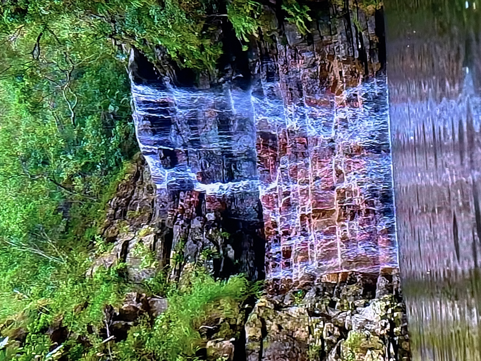The image depicts a cascading waterfall surrounded by lush greenery and rocky terrain. The water flows over several tiers of rocks, creating a textured effect as it descends into a calm pool of water at the base. The rocks themselves are layered and appear to be a mix of dark and reddish-brown hues, with patches of moss and small plants growing in the crevices. Dense foliage, primarily shades of green, covers the surrounding cliffs and upper edges of the waterfall. The water reflects the colors of the rocks and the surrounding vegetation, with hints of blue and purple visible. The overall impression is a natural, serene landscape. There is no discernible evidence to suggest this scene is located in the city of Krasnokamsk, Russia.