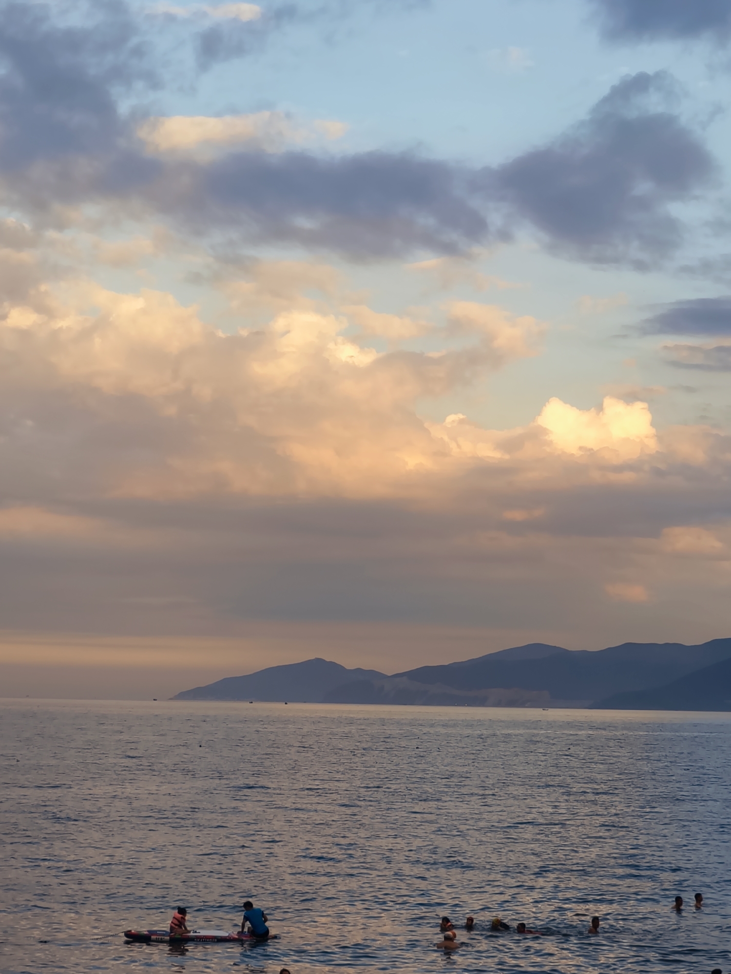 The image depicts a serene sunset scene at a beach in Nha Trang, Vietnam. The sky is a canvas of soft pastels, with hues of orange, pink, and blue dominating the clouds. In the foreground, the calm sea water reflects the gentle colors of the sky. Several people are enjoying the water; some are swimming, while others are on a paddleboard. A paddleboard with two individuals, one wearing a life vest, is positioned in the lower left quadrant. In the distance, a mountainous silhouette lines the horizon, adding depth to the tranquil seascape. A few small boats are visible on the water near the mountains. The overall atmosphere is peaceful and idyllic, characteristic of a late afternoon at a tropical beach.