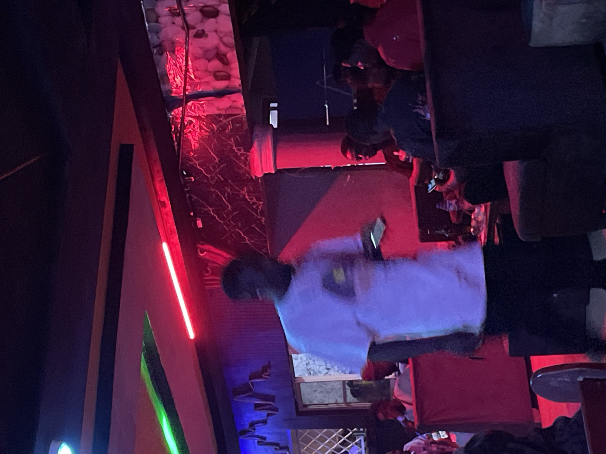 The image depicts an indoor scene at a club in Durumi, Nigeria. The environment is dimly lit, illuminated by vibrant red and green neon lights and some ambient lighting. In the foreground, a person in a white t-shirt is partially visible, appearing to be walking or standing. Several other individuals are present throughout the club, some seated and others standing. They are dressed casually. The club's interior features seating areas, possibly booths and chairs, and a bar or counter area. There are decorative elements on the walls, including what appears to be textured wallpaper or panels. Bottles and glasses are visible on tables, suggesting a bar or lounge setting. The overall atmosphere suggests a social gathering or nightlife scene.