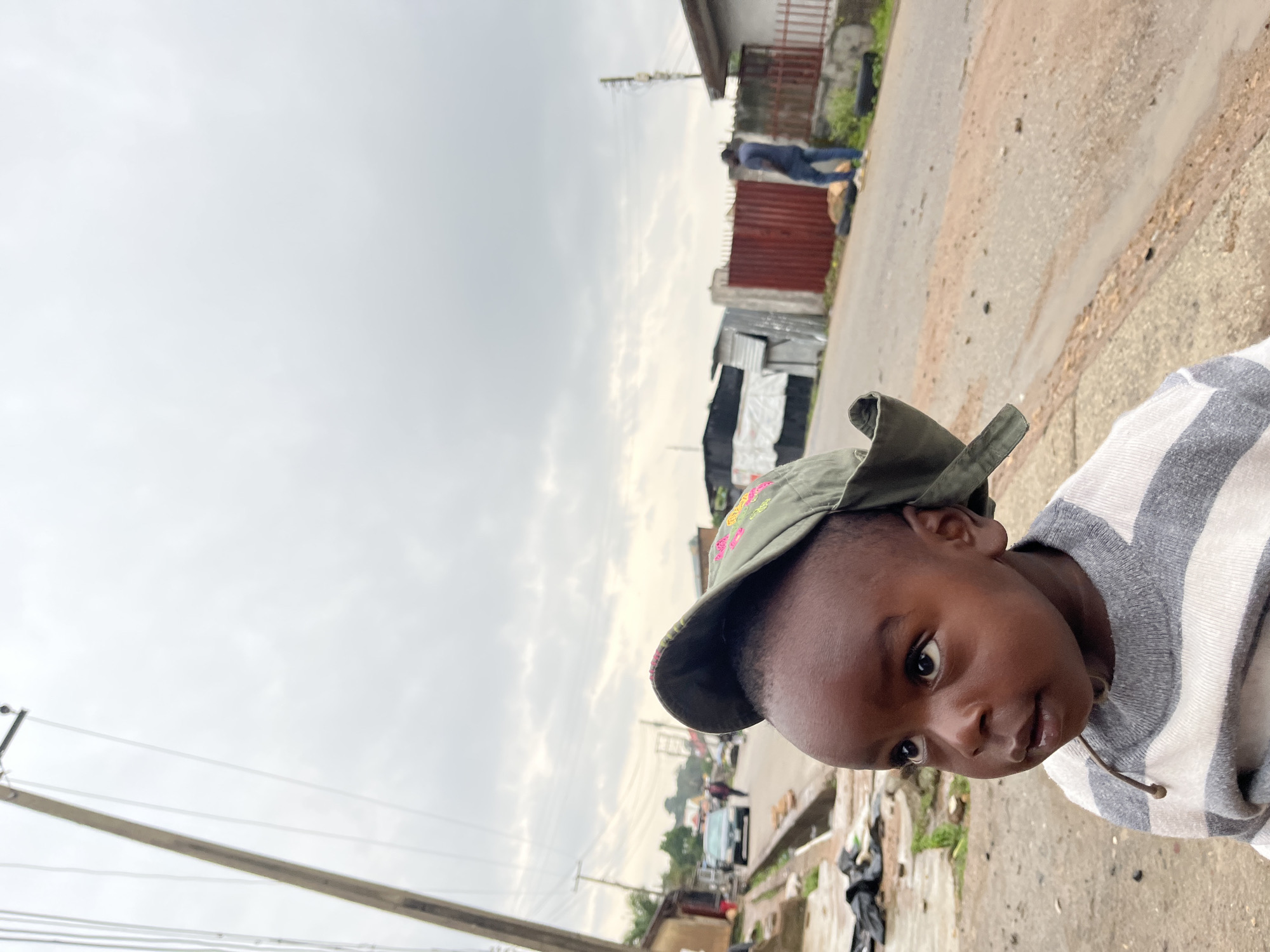 This is a close-up, portrait-style shot of a young child in Jos, Nigeria. The child is wearing a green baseball cap with embroidered floral designs and a grey and white striped sweater. The child's face is turned towards the camera, with dark eyes and a neutral expression.
The background is blurred, but appears to be an outdoor scene with a dirt road, some buildings with corrugated metal roofs, and a red metal fence. A few other people and vehicles are visible in the distance, suggesting a street or a market area. The sky is overcast, indicating a cloudy or potentially rainy day. The overall lighting suggests it is daytime. The ground appears to be a mix of dirt and pavement with some debris scattered around. The image is taken from a slightly low angle, looking up at the child. There is no visible text in the image.