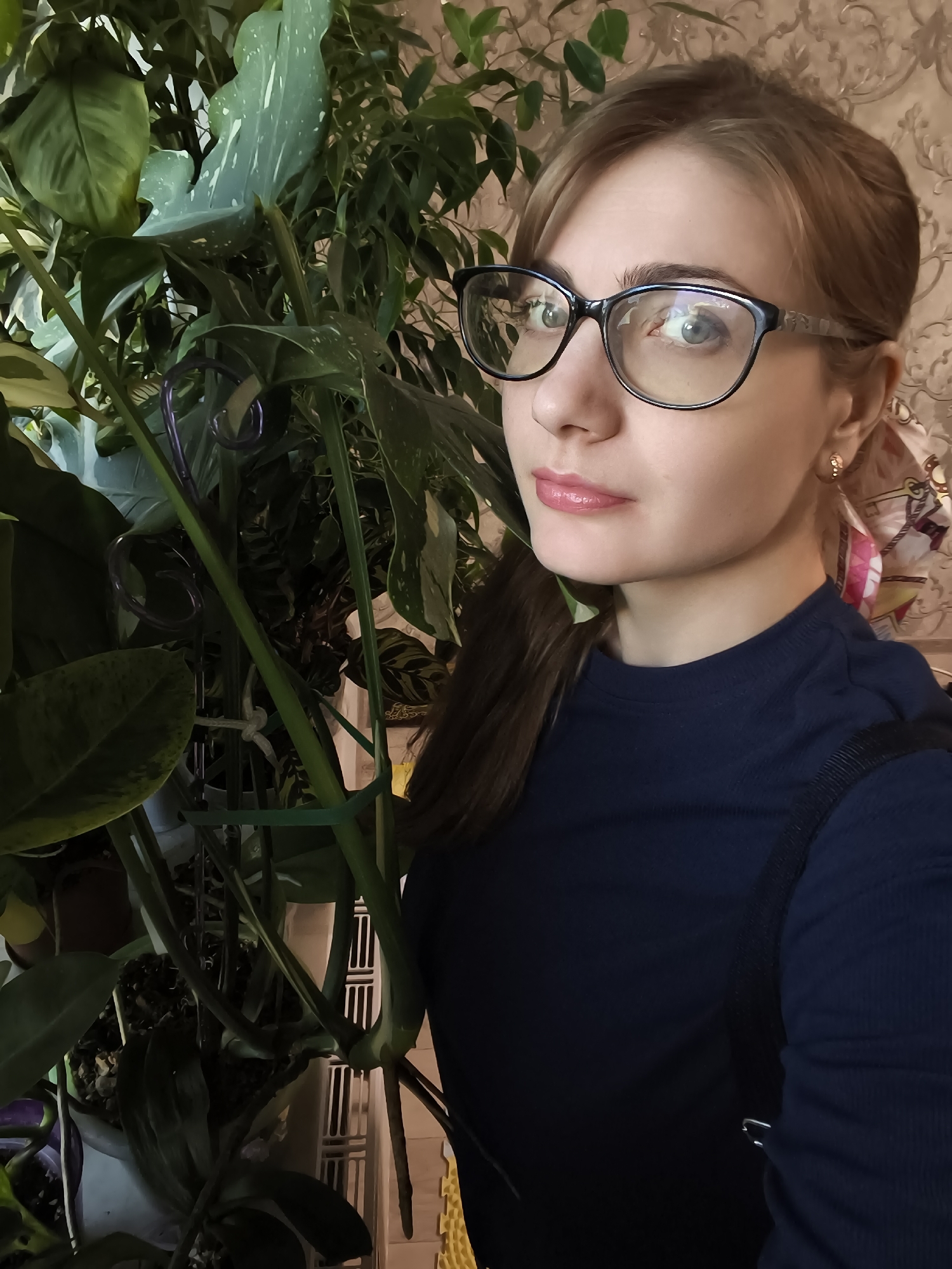 In a medium shot, a young woman with light brown hair pulled back into a ponytail and wearing black-rimmed glasses looks to her right, slightly away from the camera. Her makeup is subtle, with emphasis on her pink lipstick and mascara. She has on a dark navy blue long-sleeved shirt.
She is positioned to the right of a large assortment of indoor plants, which fill the left and center of the frame. The plants have large, glossy green leaves, some with distinctive white variegation. One of the plants is supported by a green stake and ties. The background shows a textured wallpaper with a light brown floral pattern.
The lighting suggests an indoor setting, likely during the daytime given the brightness. The woman's expression is neutral, perhaps a little thoughtful. There is no visible text in the image. The scene appears to be a home environment in Podkletnoe, Russia, where the woman is tending to or admiring her plants.
