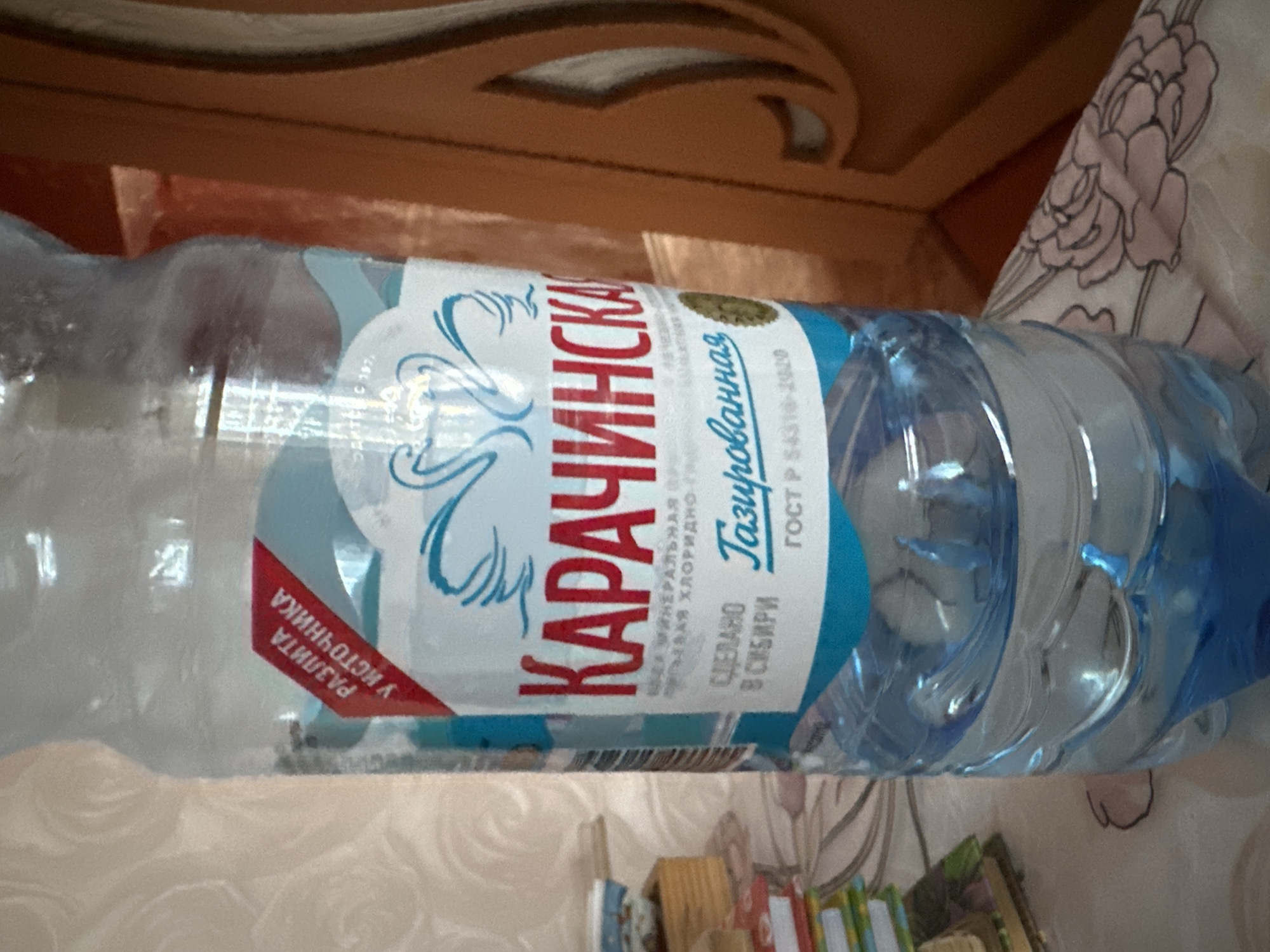 A close-up shows a plastic bottle of "Karachinskaya" sparkling mineral water. The label indicates the water is bottled at the source and produced in Siberia, according to GOST R 54316-2020 standards. The surrounding environment includes what appears to be bedding with a floral pattern and a wooden surface, possibly furniture. The presence of books or other small objects is also indicated at the bottom of the frame. The specific location within the city of Sayansk, Russia, cannot be determined from the image alone.