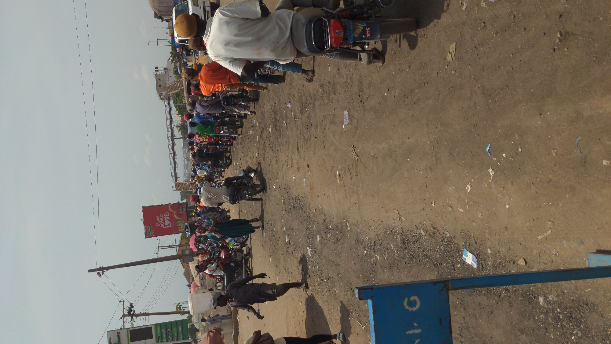 This daytime image captures a bustling street scene, likely in Ado, Nigeria, viewed from a slightly elevated perspective. The ground is an unpaved, dusty road, marked with tire tracks and scattered debris like plastic and paper. The sky is bright but hazy, with utility poles and electrical wires crisscrossing the upper left.
Numerous people are present, predominantly men, mostly on motorcycles, which appear to be either stalled in traffic or waiting in a queue. A man in a light-colored shirt and a brown hat sits on a motorcycle in the foreground, facing away from the viewer. Another man in a dark shirt and cap walks across the dusty ground towards the right. Several pedestrians are scattered throughout the scene, including a woman in a green dress walking with an umbrella. Many individuals on motorcycles wear helmets, and some carry umbrellas, suggesting sunny and possibly hot weather.
Prominent objects include a large red billboard in the mid-ground on the left, displaying the text "Show Some Love" with an associated image. Below it, a green sign, possibly for a gas station, is partially visible. In the foreground on the right, a blue structure features white, partially legible text, which could be "G.1" or "9.1". In the background, buildings are visible, and a large white vehicle, possibly a bus or van, is partially seen on the far left. The overall scene depicts a typical busy day on a dirt road in a populated urban or peri-urban area, characterized by motorcycle traffic and pedestrian activity.