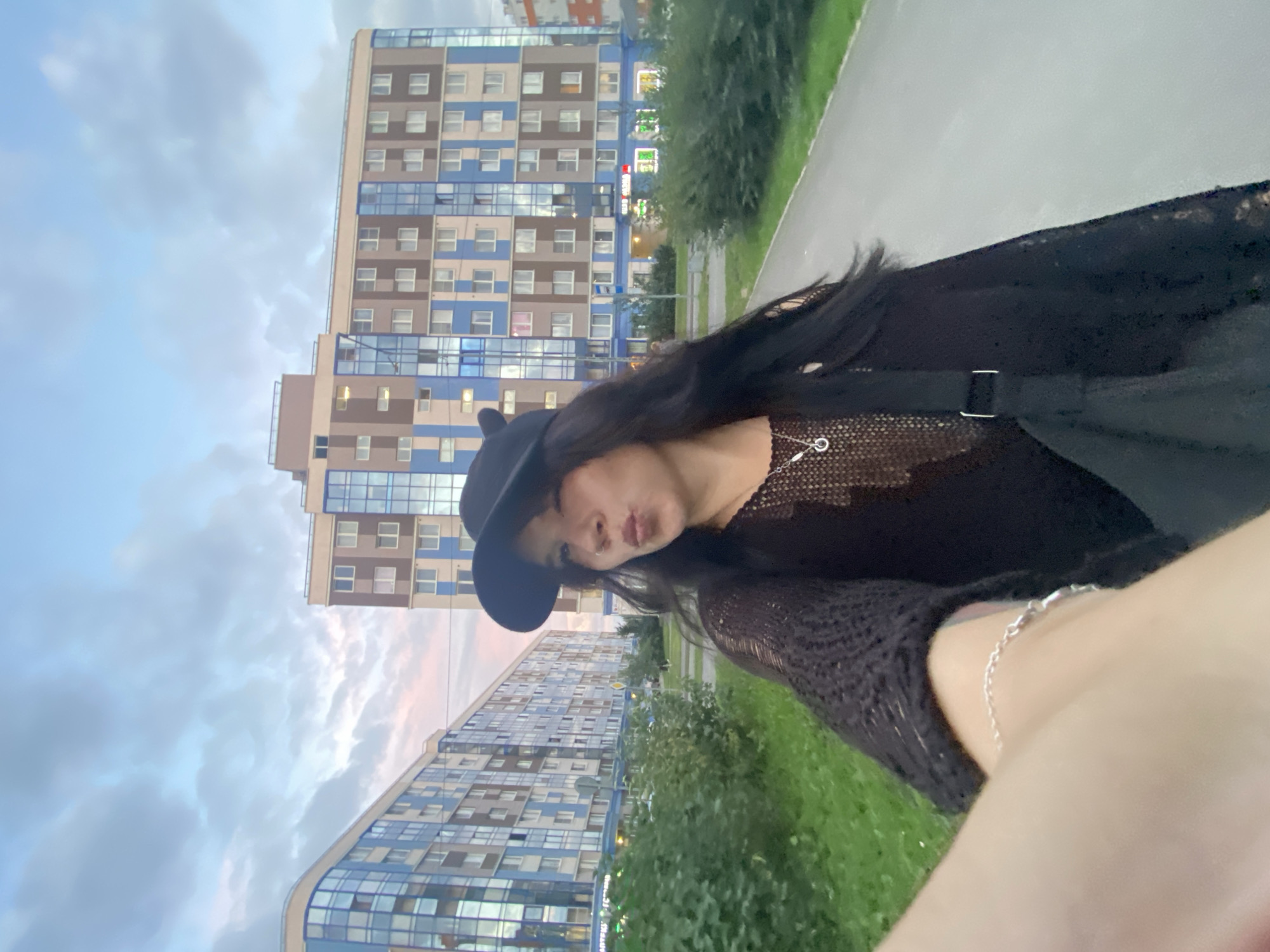 A selfie is taken by a young woman with long, dark hair, wearing a black baseball cap, a black mesh top with a silver necklace, and a black crossbody bag. She is standing on a paved pathway in front of several multi-story residential buildings with blue and brown facades. The sky is partly cloudy, with a hint of pink from the sunset. Green grass and bushes line the pathway. The buildings appear to be located in the city of Yanino-1, Russia, as indicated by the architectural style and potential signage visible on the buildings. The woman is making a kissy face at the camera, and her right arm is extended to hold the phone for the photo. A silver bracelet is visible on her left wrist.