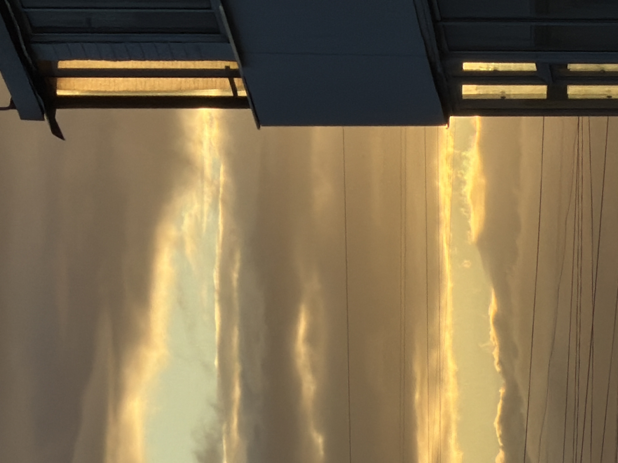 The image captures a building facade against a sky filled with clouds illuminated by golden sunlight, likely during sunset or sunrise. The building is a multi-story structure with visible windows, some of which are lit from within, casting warm yellow light. The sky is a mix of dark grey and lighter, golden hues, indicating a dramatic interplay of light and shadow in the clouds. Several horizontal lines, likely power or telephone wires, stretch across the lower portion of the frame. The setting appears to be urban, given the presence of the building and wires. A specific location in Khimki, Russia is mentioned, suggesting this scene is a part of that city's landscape, possibly a residential or commercial building. No people or specific actions are depicted.