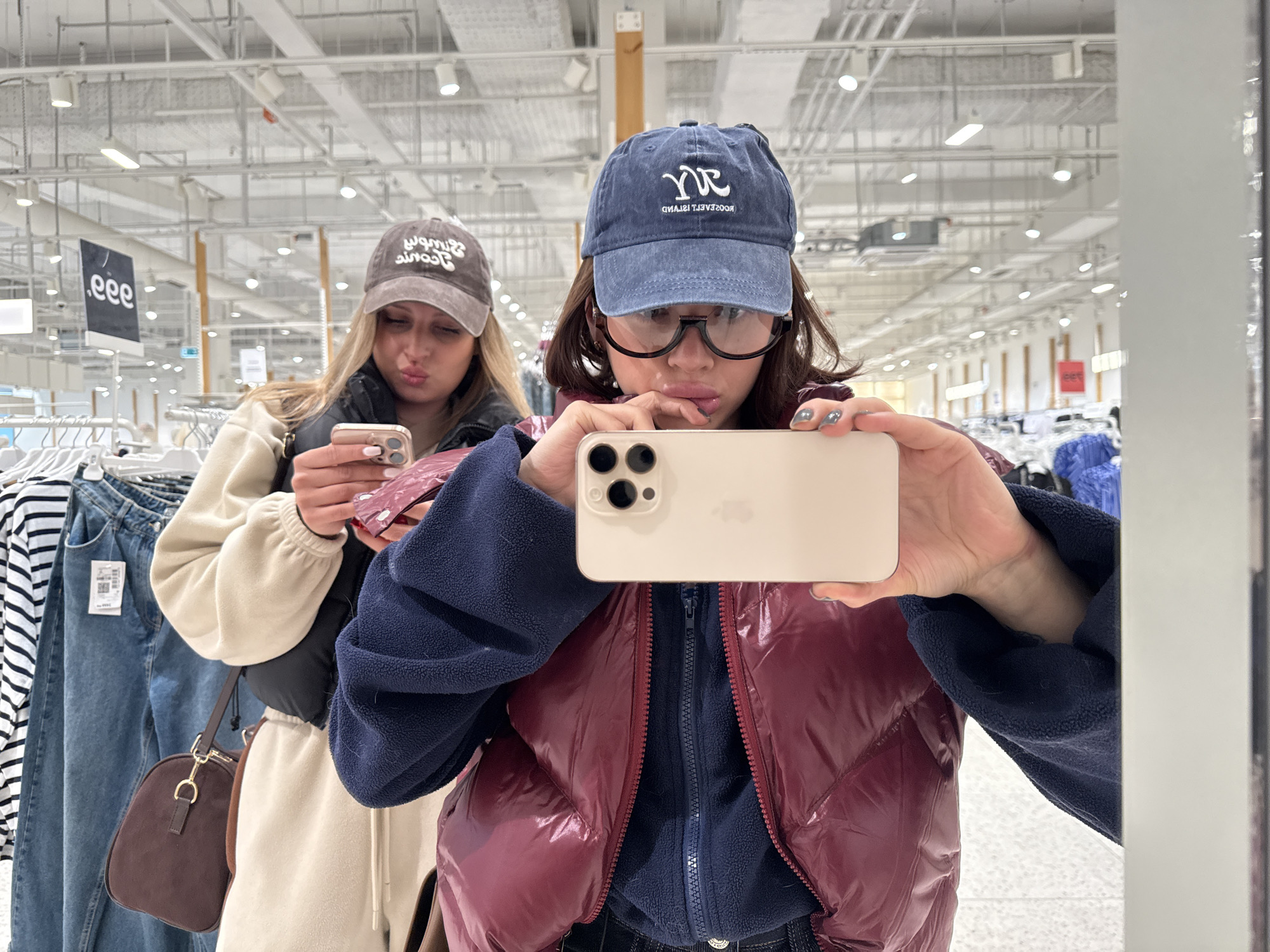This media file captures two young women in what appears to be a brightly lit retail clothing store, likely in Perm, Russia. The image is a mirror selfie taken by the woman in the foreground.
The foreground subject wears a navy blue baseball cap with "YR" (reversed) and "ROOSEVELT ISLAND" (reversed) embroidered on it. She also has large, clear-framed glasses, a dark blue fleece jacket under a maroon puffy vest. She holds a light-colored iPhone (identifiable by its triple-lens camera setup) horizontally, capturing her reflection. Her fingernails are painted grey, and she has a slightly pouting expression.
Behind her, reflected in the mirror, is another woman wearing a brown baseball cap with reversed, unreadable text, a black vest over a cream or beige long-sleeved top, and matching cream sweatpants. She also holds a light-colored iPhone and has a similar pouting expression. A small, dark brown shoulder bag with gold hardware is visible on her side.
The setting is a typical clothing store with rows of merchandise visible on racks, including blue jeans and striped tops. The ceiling features numerous overhead fluorescent lights and exposed ventilation ducts, characteristic of a large retail space. In the background, a black sign with ".eee" (reversed) is visible, likely part of a promotional display. The overall ambiance is casual, suggesting a shopping trip.