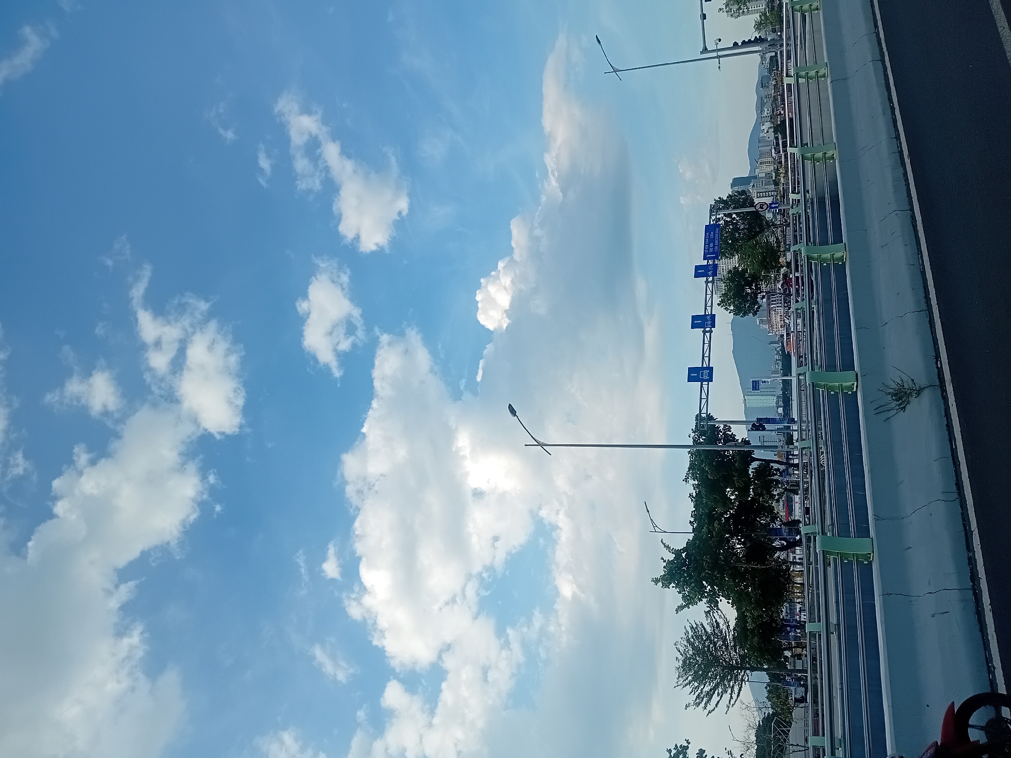 A wide shot of a modern highway overpass in the city of Da Nang, Vietnam, under a partly cloudy sky. The overpass consists of multiple lanes, separated by concrete barriers and metal guardrails, with green metal posts supporting the guardrails. Trees line the sides of the overpass, and beyond them, the city skyline is visible, featuring tall buildings and a distant mountain range. Road signs are mounted on overhead gantries, indicating directions and traffic regulations. The foreground shows a portion of the asphalt road and a red motorcycle. The overall scene suggests a vibrant urban environment with well-developed infrastructure.